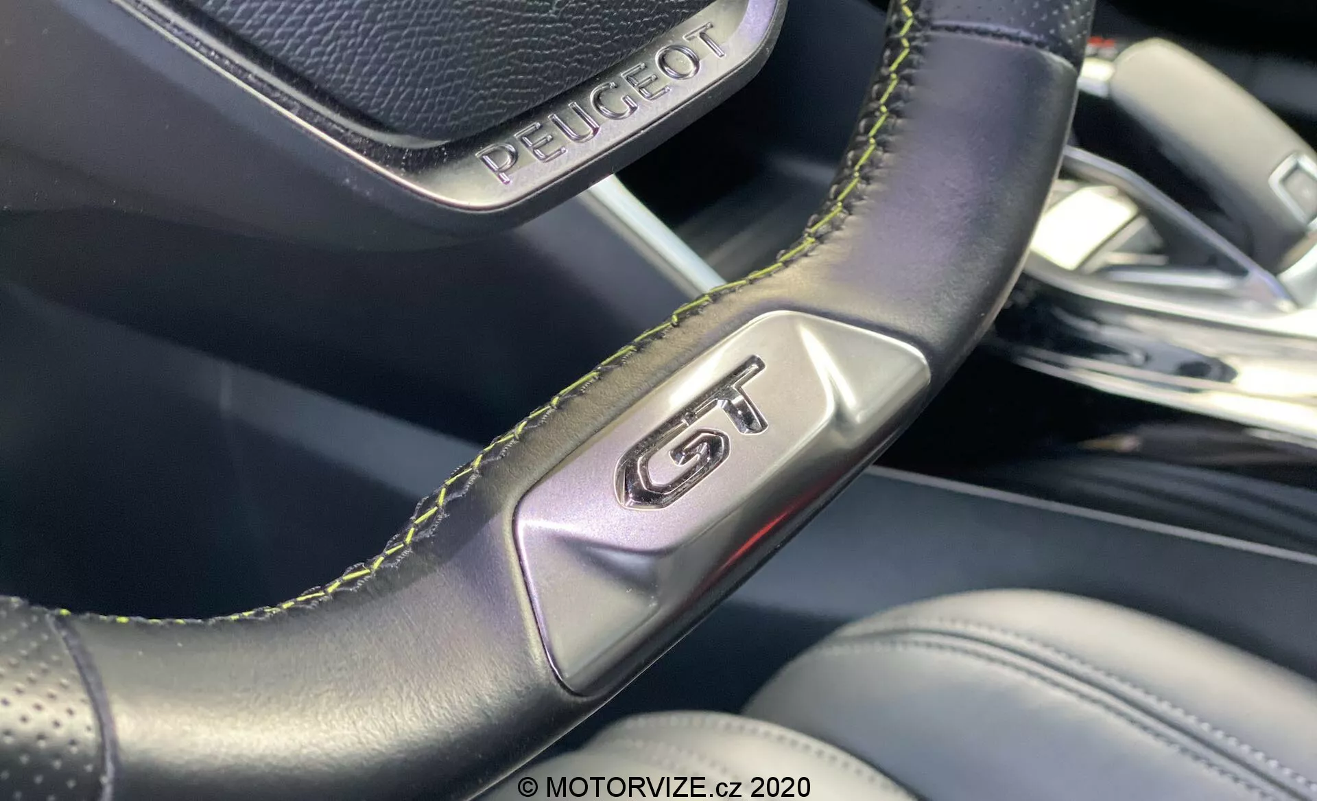 Blízky záber na interiér vozidla Peugeot 2008 2020, zvýrazňujúci znak 'GT' na perforovanom koženom volante so zeleným prešívaním, zdôrazňujúci športovosť a luxus. Obraz sa zameriava na detaily remeselného spracovania a rozmazané pozadie odhaľuje prémiové materiály interiéru a moderný dizajn.