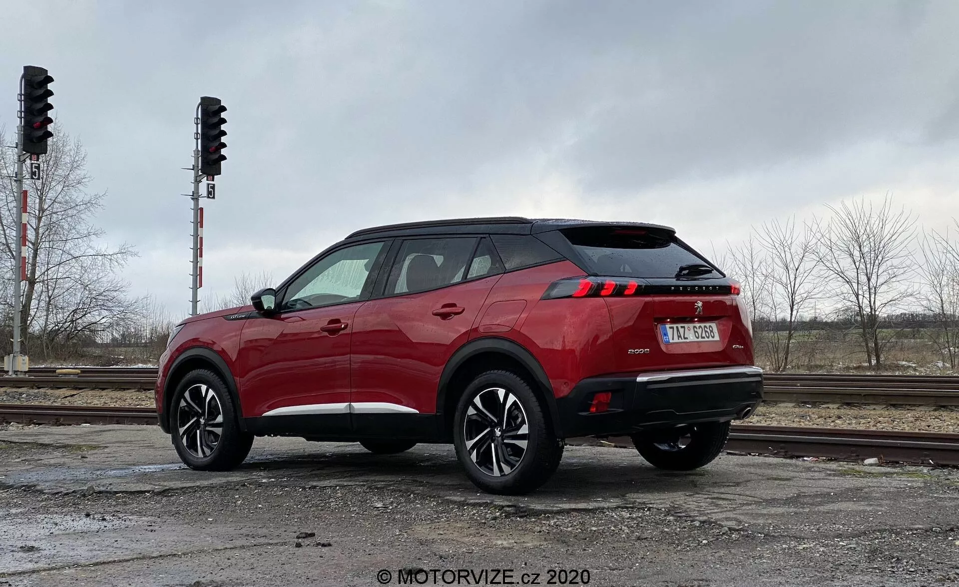 Obrázok Peugeot 2008 2020 (Peugeot 2008 1.2 PureTech 100 Active) v žiarivej červenej farbe, ktorý zobrazuje moderné SUV umiestnené vonku blízko železničných koľají a výstražných signálov. Snímka zhotovená zo zadného trojštvrťového pohľadu ukazuje pravú stranu vozidla s elegantnými LED zadnými svetlami, čiernym plastovým obložením okolo oblúkov kolies a dolných prahov, viaclúčovými zliatinovými kolesami s nízkoprofilovými pneumatikami a športovou charakterovou líniou na dverách. Záber z úrovne zeme zvýrazňuje pôsobivý postoj a výšku SUV.