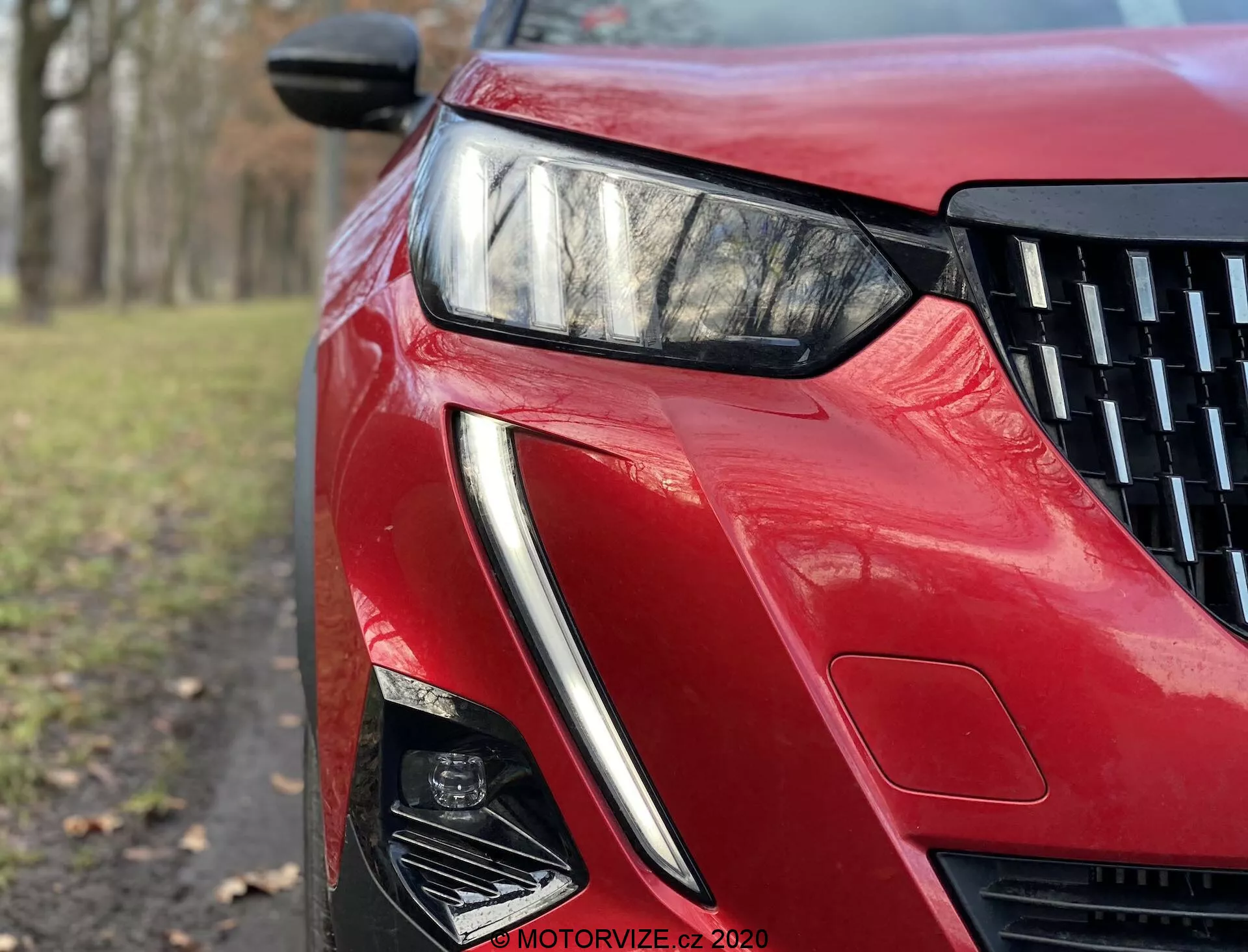 Detailný pohľad z nízkeho uhla na prednú ľavú časť modelu Peugeot 2008 2020 s lesklým červeným lakom. Obrázok zdôrazňuje elegantný, uhlový LED svetlomet, ostrý dizajn nasávania vzduchu a tvarované karosárske línie, odrážajúce pokročilé osvetľovacie technológie a aerodynamiku.