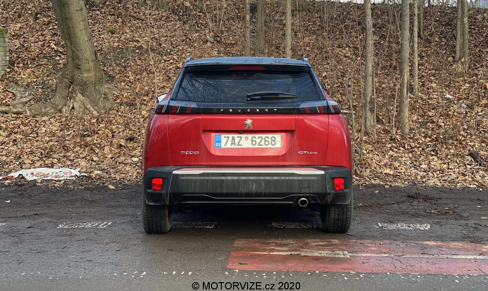 Zadný pohľad na červené auto zaparkované vonku na pozadí bezlistých kríkov alebo stromov, čo naznačuje jeseň alebo zimu. Medzi vlastnosti auta patrí centrálny znak a štítok na veku kufra, registračná značka, zadný stierač, zadný spojler, moderné zadné svetlá, odrazky a zadné parkovacie senzory na nárazníku, výrazný zadný nárazník s viditeľnou výfukovou rúrou, zadná ochranná platňa, anténa v štýle žraločej plutvy na streche a olupujúca sa nálepka na ľavej strane spodnej časti veka kufra.