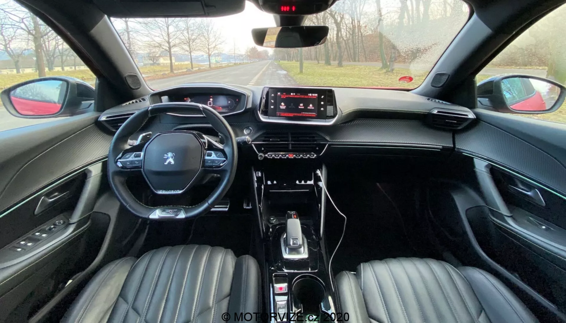 Interiérový záber na Peugeot 2008 2020 (peugeot-2008-1-2-puretech-100-active) z pohľadu zadného sedadla. Obrázok ukazuje predné sedadlá potiahnuté čiernou kožou s ryhovaným dizajnom, integrovanými opierkami hlavy, multifunkčným volantom s logom Peugeot a modernou stredovou konzolou. Palubná doska obsahuje dotykovú obrazovku infotainmentu s minimom fyzických ovládacích prvkov, čo naznačuje orientáciu na ovládanie dotykom. Auto obsahuje automatickú radiacu páku, voliče jazdných režimov, elektronickú parkovaciu brzdu a úložné priehradky. Atmosféra naznačuje jasný deň s jasným výhľadom na cestu vpredu.