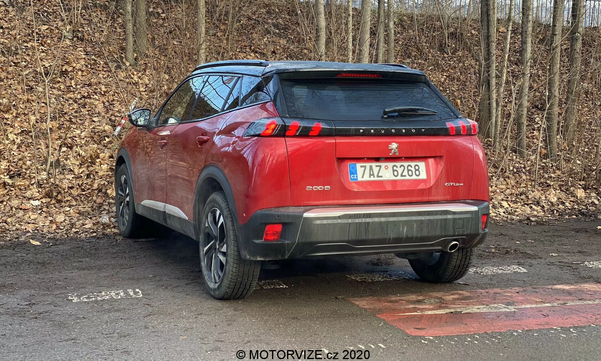 Táto fotografia zobrazuje zadný pohľad na červené SUV Peugeot 2008 2020 (Peugeot-2008-1-2-PureTech-100-Active) zaparkované vonku. Obraz zo zadného trojštvrtinového uhla ukazuje zadný a bočný profil auta, s dôrazom na prvky ako sú zadné svetlá, zadný nárazník s možnými parkovacími senzormi, strešný spojler a čiastočný dizajn kolies. Moderné LED zadné svetlá, dvojfarebné lakovanie, centrálne umiestnená poznávacia značka a reflexné prvky na spodnej časti nárazníka zvyšujú jeho bezpečnosť a estetický dojem. Fotografia bola urobená na mokrom, listami pokrytom povrchu s pozadím stromov, čo naznačuje mimo mestské prostredie. Zachytené sú jemné detaily ako textúra materiálov, priehľadnosť skla a vzor behúňa pneumatiky.
