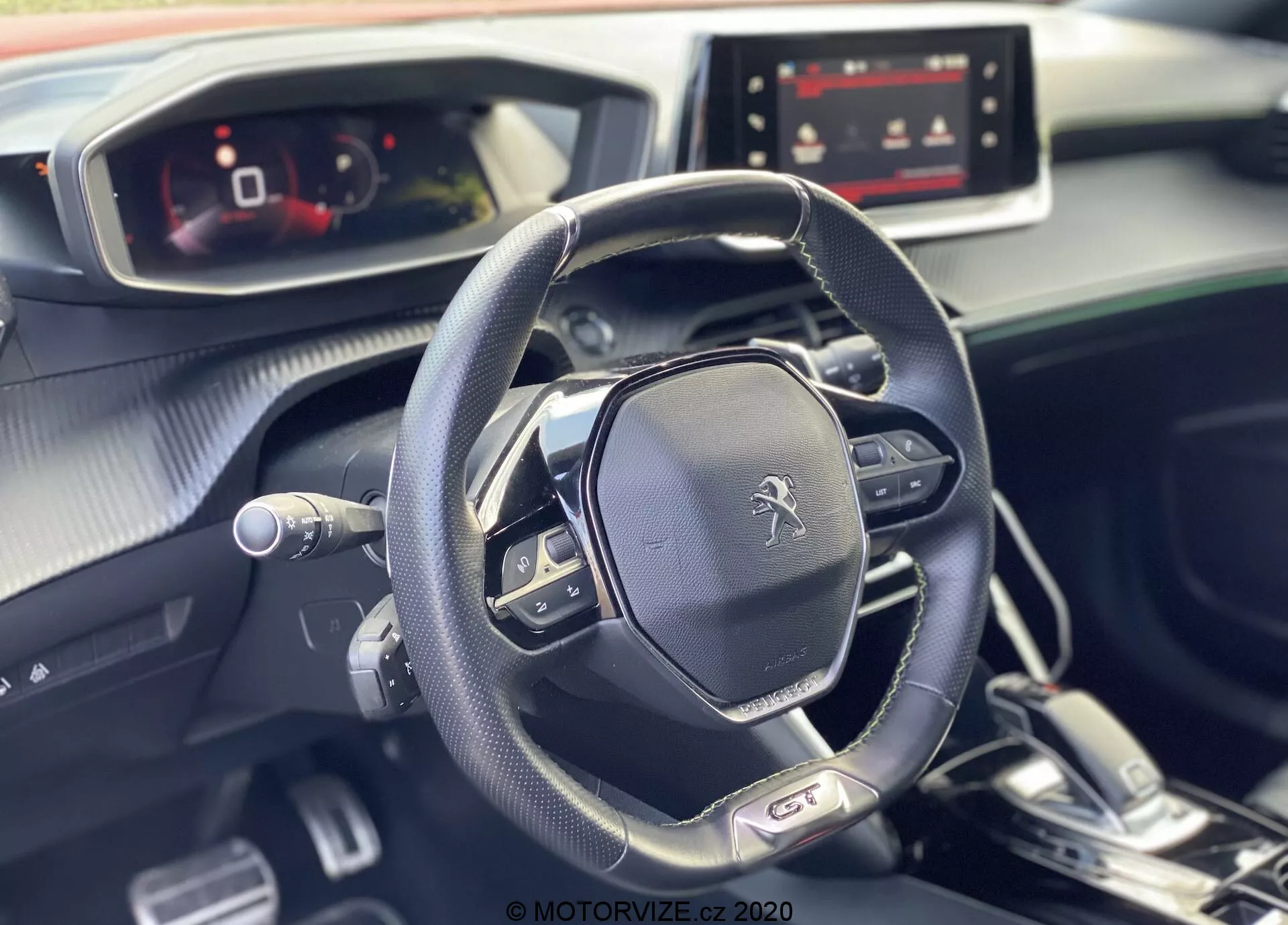 Obráz zobrazuje moderný a elegantný interiér vozidla Peugeot 2008 2020, so zameraním na multifunkčný volant a plne digitálny prístrojový panel z pohľadu pasažiera. Na volante je výrazný znak Peugeot, ktorý obsahuje ovládanie médií, nastavenia tempomatu a radiace páčky, čo naznačuje automatickú prevodovku s možnosťou manuálneho radenia. Palubná doska, navrhnutá s kombináciou mäkkých dotykových materiálov a kovových akcentov, obsahuje zapnutú obrazovku infotainmentu, ktorá naznačuje rôzne možnosti pripojenia a navigácie. Prírodné denné svetlo zvyšuje bezchybnú a príjemnú atmosféru interiéru vozidla.