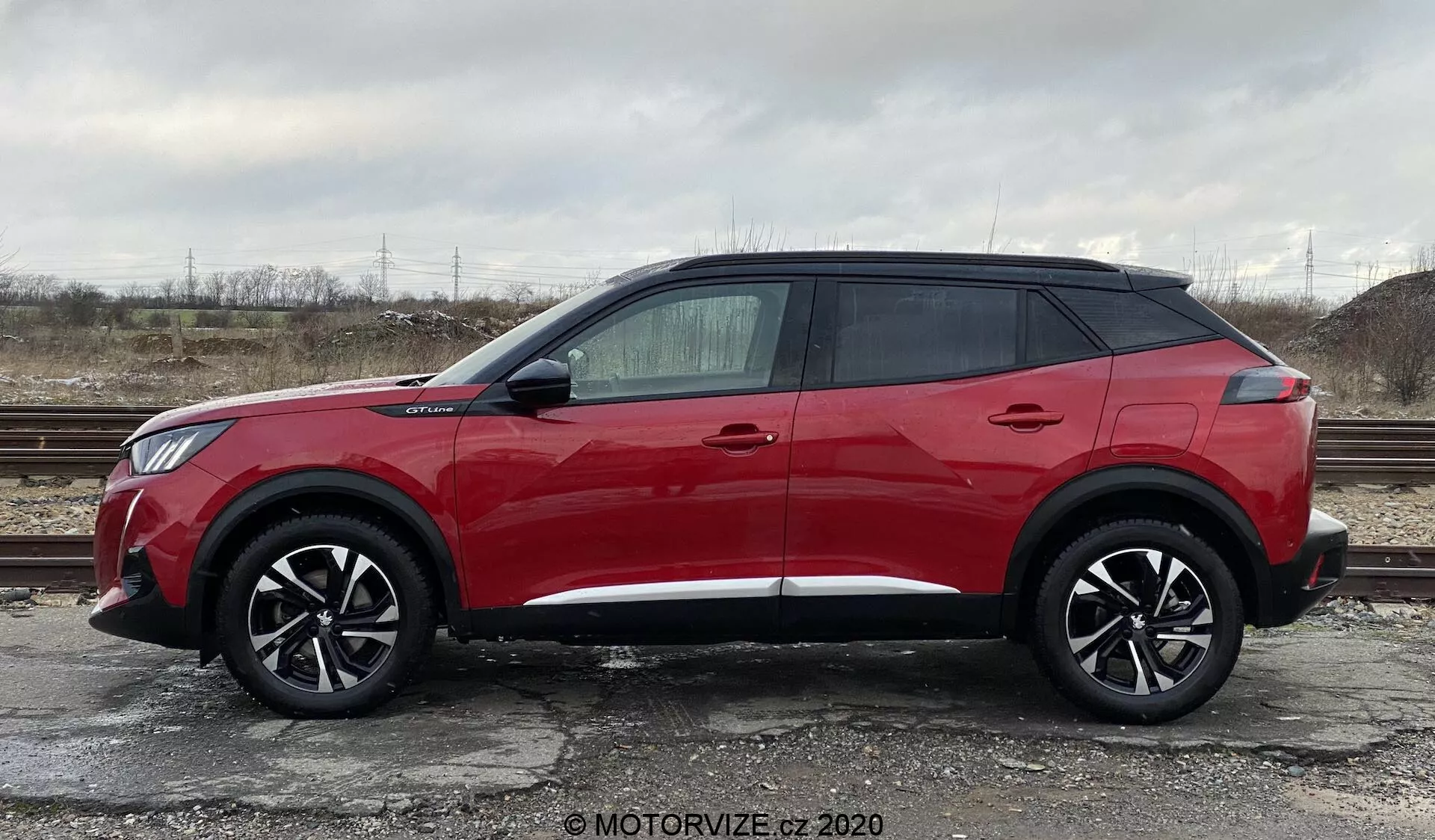 Bočný profil jasne červeného Peugeot 2008 2020 SUV, model 1.2 PureTech 100 Active, zaparkovaného na štrkovom povrchu so sporou vegetáciou a zamračenou oblohou v pozadí. Na obrázku je vozidlo s veľkými čiernymi viaclúčovými zliatinovými kolesami, elegantným súčasným dizajnom s vysokou bočnou líniou, štyrmi kľučkami na dverách, tónovanými sklami, vystupujúcimi bočnými zrkadlami s integrovanými smerovkami a plávajúcim strešným dizajnom s čiernymi zadnými stĺpikmi. Podlžná ozdobná línia pozdĺž spodnej časti vozidla.
