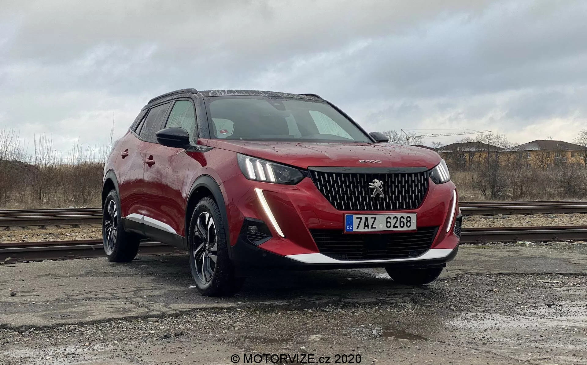 Červené SUV Peugeot 2008 2020 (Peugeot-2008-1-2-PureTech-100-Active) zaparkované vonku proti zatiahnutej oblohe so suchou pôdou a stavebnými sutmi v pozadí. Vozidlo je zachytené z nízkeho a blízkeho predného trojštvrťového uhla, čo ukazuje prednú mriežku s logom Peugeot, elegantné LED svetlomety, hmlovky, viaclúčové zliatinové kolesá a metalický červený lak s čiernym plastovým obložením okolo blatníkov. Bočné zrkadlá, čiastočne viditeľné, sa zdajú byť vo farbe karosérie s integrovanými smerovkami a šikmé čelné sklo zaisťuje dobrú viditeľnosť pre vodiča. Pravdepodobné funkcie zahŕňajú parkovacie senzory a kamery, čo možno usúdiť z jeho moderného dizajnu.