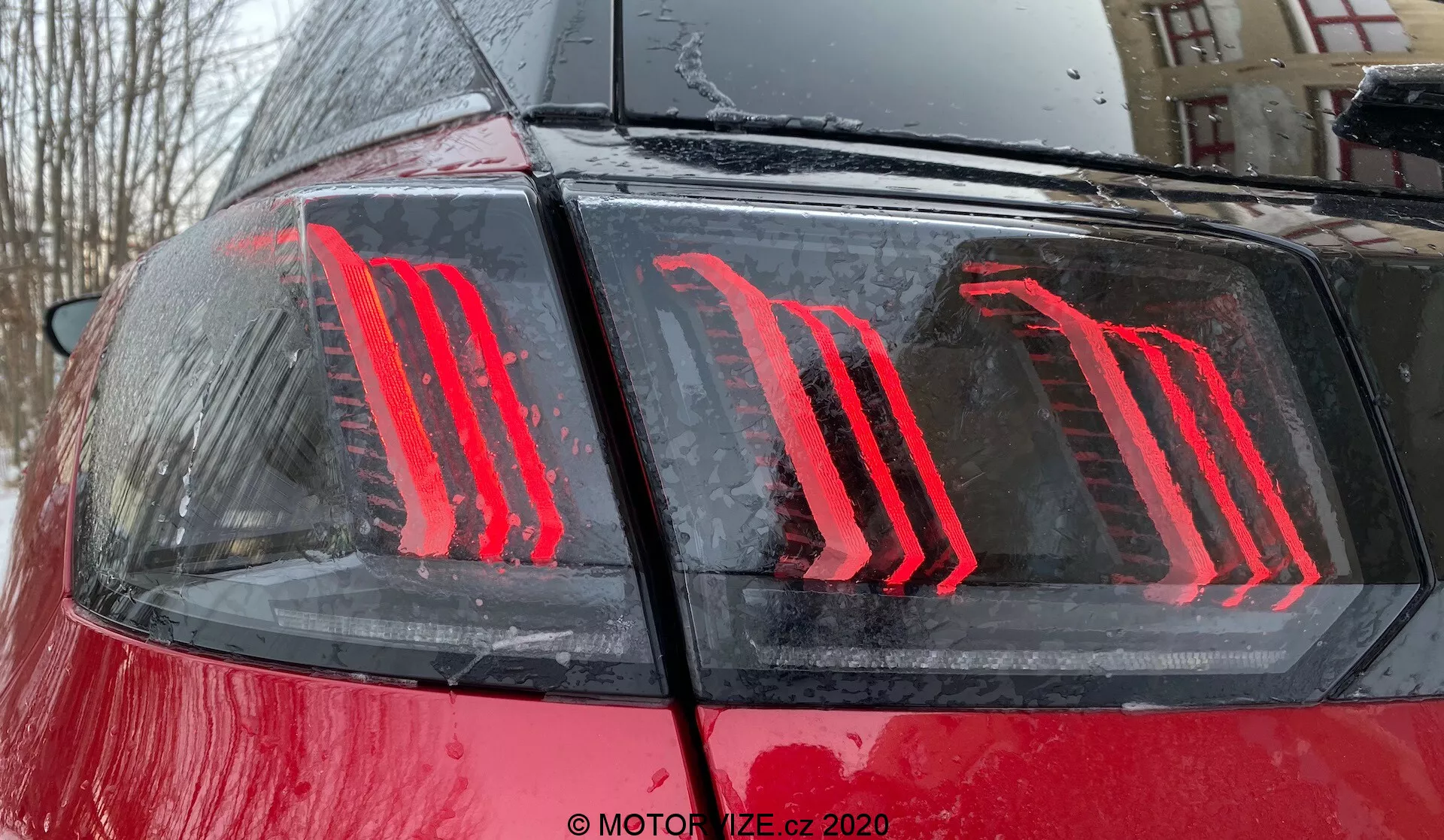Detailný záber na zadnú časť auta Peugeot 3008 z roku 2021, zdôrazňujúci moderný vertikálne osvetlený dizajn LED zadného svetla na lesklej červenej karosérii s kvapkami vody, naznačujúc vonkajšie prostredie počas alebo po daždi; zadné svetlo obsahuje energeticky úspornú technológiu a slúži ako brzdové a denné svetlo.