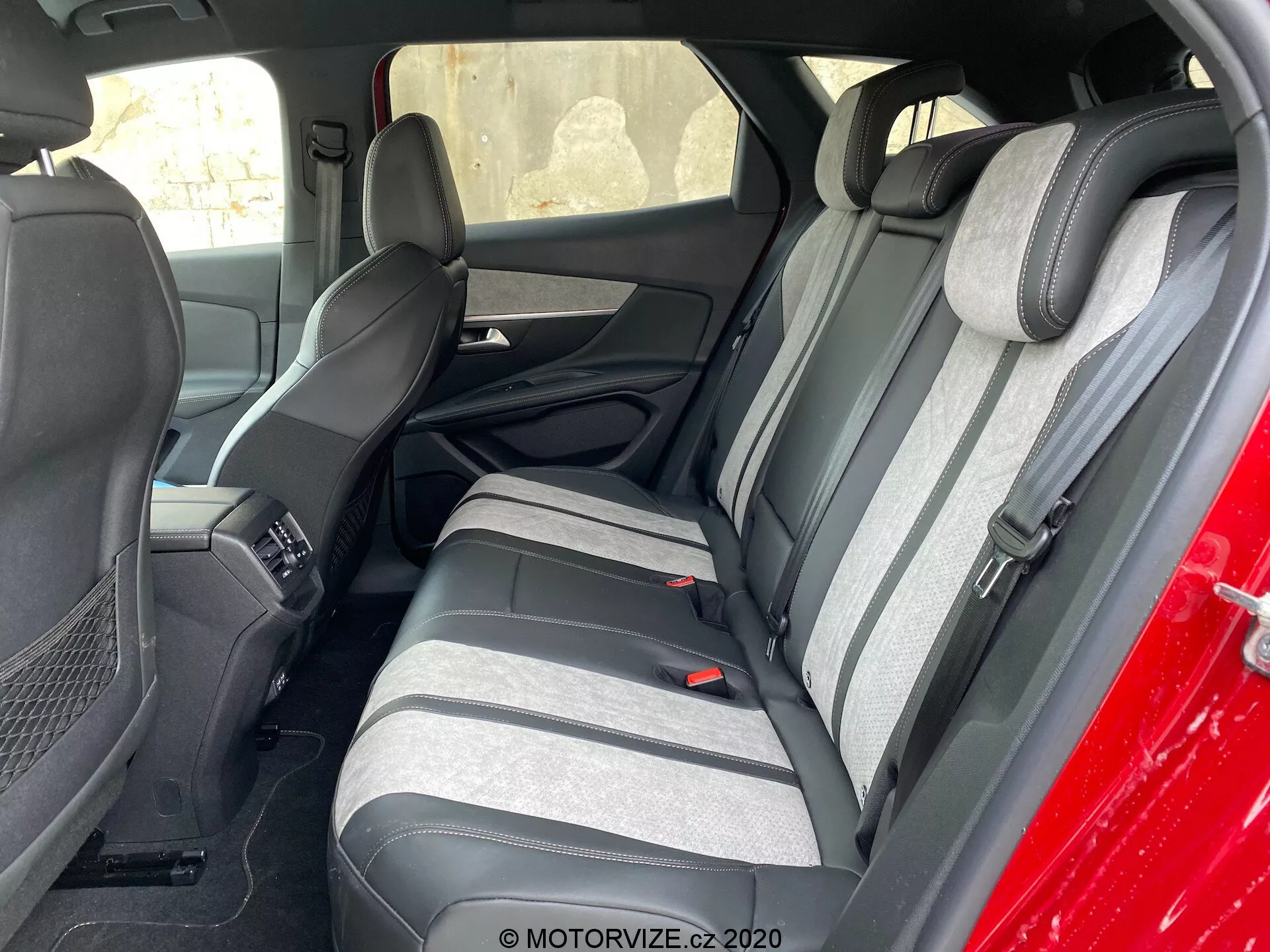 Interiérový záber vozidla Peugeot 3008 z roku 2021 zameraný na zadnú sedadlovú časť, viditeľný zvonku, pod uhlom k vodičovej strane. Zadné sedadlá sú potiahnuté dvojfarebnou tkaninou v tmavých a svetlých odtieňoch sivej s textúrovaným pásovým vzorom. Viditeľné sú aj zodpovedajúce zdvihnuté opierky hlavy. Viditeľná je tiež časť predných sedadiel a stredovej konzoly. Zadné sedadlá majú trojbodové bezpečnostné pásy a opierku s integrovanými držiakmi na poháriky. Vnútorné panely dverí zodpovedajú farbám sedadiel a obsahujú mriežky reproduktorov a ovládacie prvky.