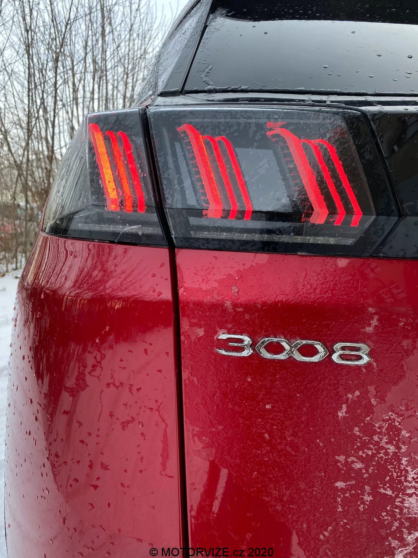 Detailný pohľad na zadnú časť červeného Peugeot 3008 2021 z nízkeho uhla, ukazujúc charakteristické tri vertikálne LED svetlá vytvárajúce vzor pazúrov a modelové označenie '3008'. Mokrá povrchová úprava auta, pokrytá kvapkami vody a nečistotami alebo snehom, zdôrazňuje nedávne použitie v nepriaznivom počasí za podmienok oblačného osvetlenia.