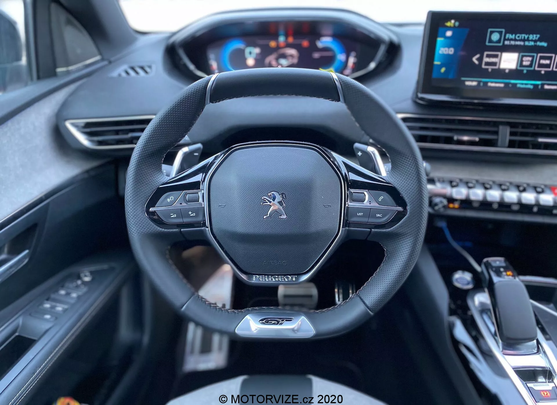 Pohľad na interiér z miesta vodiča vozidla Peugeot 3008 z roku 2021, zobrazujúci volant a palubnú dosku. Obrázok sa sústreďuje na centrálny volant, ktorý má logo Peugeot, moderný dizajn s plochým spodkom, koženým okrajom a zabudovanými ovládačmi pravdepodobne pre nastavenia zvuku a tempomatu. Za volantom sú viditeľné páčky pre manuálne radenie. Digitálny prístrojový panel zobrazuje tachometer a otáčkomer, osvetlený, čo signalizuje, že vozidlo je zapnuté. Na pravej strane volantu je čiastočne viditeľný centrálny dotykový infotainment systém a rôzne ovládacie tlačidlá, spolu s radiacou pákou a stredovou opierkou v dolnom popredí, čo naznačuje moderné, technologicky vyspelé vozidlo s luxusným interiérom.