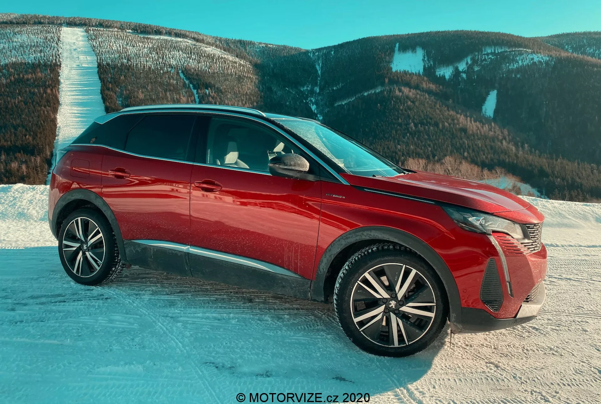 SUV Peugeot 3008 z roku 2021 v červenej farbe, fotografovaný z pohľadu tri štvrtiny spredu na zasneženom povrchu s horskou krajinou v pozadí, s dôrazom na pokročilé LED svetlomety, prepracované hliníkové kolesá, lesklý povrch, aktívne mriežky chladiča a bočné zrkadlá s integrovanými ukazovateľmi smeru, zdôrazňujúci dizajnové prvky a vhodnosť pre náročné podmienky.