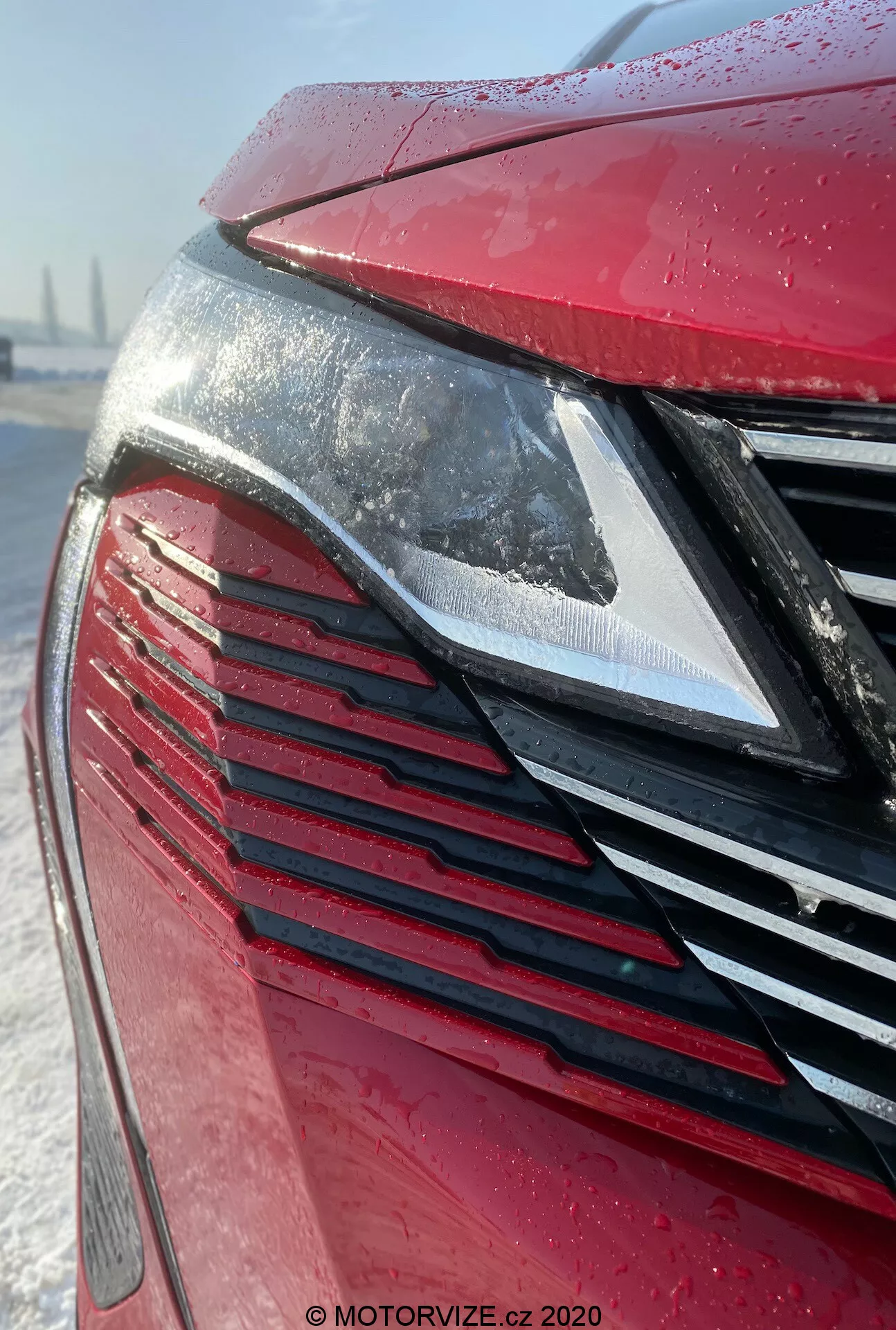 Detailný pohľad na prednú časť vozidla Peugeot 3008 2021 so zameraním na moderný LED svetlomet a mriežku s vertikálnymi lamelami v metalickej červenej farbe pokrytej kvapkami vody, čo naznačuje nedávny dážď alebo námrazu. Snehové pozadie naznačuje zimné podmienky alebo starostlivosť o auto.