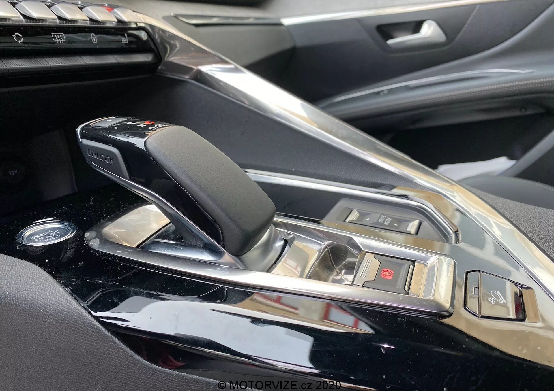 Detailný pohľad na stredovú konzolu a oblasť radiacej páky vozidla Peugeot 3008 2021 z pohľadu spolujazdca. Obsahuje elegantnú, modernú automatickú radiacu páku s koženými a chrómovými prvkami, obklopenú vysoko lesklým povrchom konzoly. Viditeľné sú tlačidlá pre rôzne funkcie vozidla a časti informačného systému, čo naznačuje luxusný interiér vozidla s vysoko kvalitnými materiálmi a sofistikovanými technologickými funkciami.