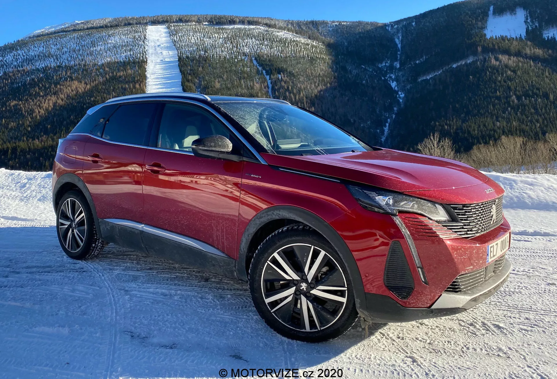 Obrázok moderného vozidla, pravdepodobne Peugeot 3008 z roku 2021, odfotografovaný z predného trojštvrťového pohľadu. Uhol zobrazuje výrazný dizajn LED svetlometov vozidla, mriežku so vzorom hrebeňa a znak na prednej strane. Predný nárazník zobrazuje sekcie prívodu vzduchu a kryty hmlových svetiel. Kapota má tvarované línie, ktoré prispievajú k aerodynamike, zatiaľ čo rozsiahle čelné sklo naznačuje dobrú viditeľnosť vpredu. Kolesá s viacerými lúčmi naznačujú športovejšiu a vyššiu úroveň výbavy. Elegantná línia strechy a dynamické línie karosérie naznačujú crossover alebo SUV. Viditeľné sú aj bočné zrkadlá s integrovanými smerovými svetlami a bočnými lištmi alebo dekoračnými prvkami. Scéna zobrazuje zasneženú cestu s horskou kulisou, čo naznačuje schopnosť vozidla zvládať rôzne terény.