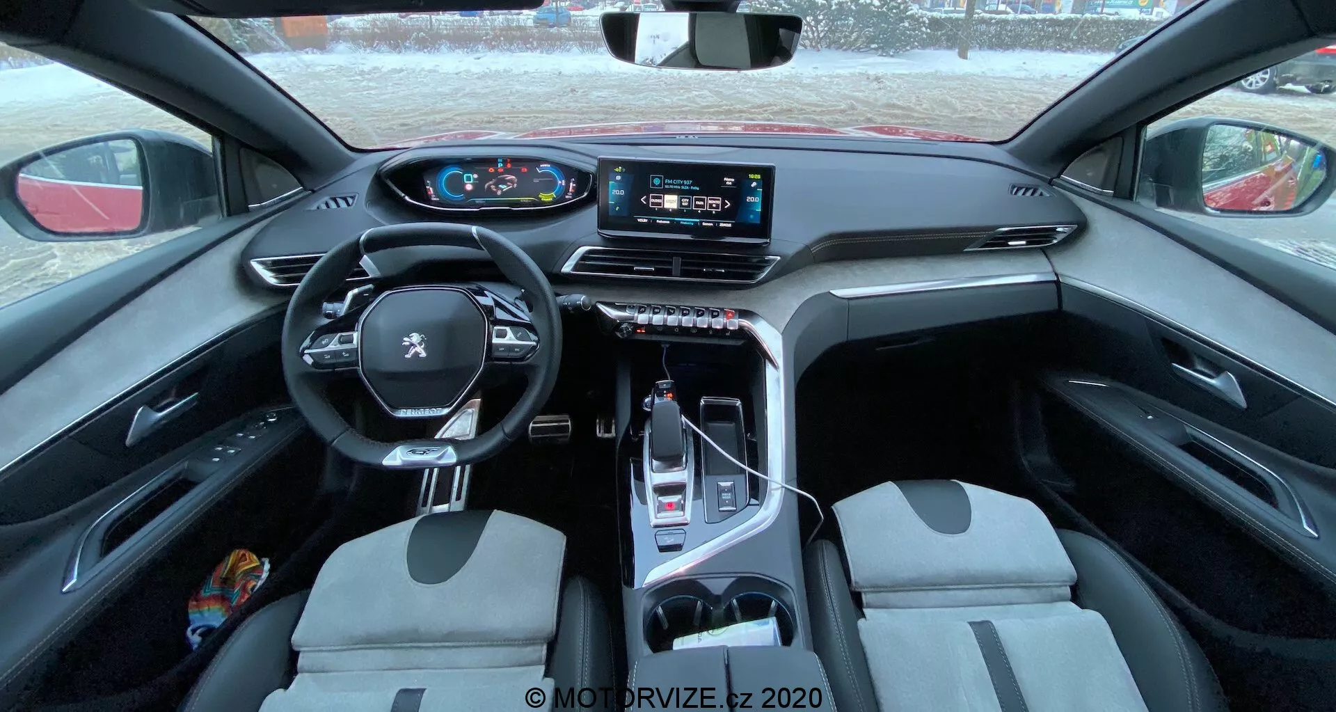 Pohľad na interiér vozidla Peugeot 3008 z roku 2021, ktorý zobrazuje multifunkčný kožený volant s namontovanými ovládačmi, digitálny prístrojový panel, veľký dotykový infotainment systém na stredovej konzole, elektronický volič rýchlostí, predné sedadlá s dvojfarebným dizajnom a zmes praktických a luxusných materiálov. Záber je urobený zo zadného sedadla, poskytujúci rozsiahly pohľad na kabínu, zdôrazňujúc dizajn a technologické funkcie.