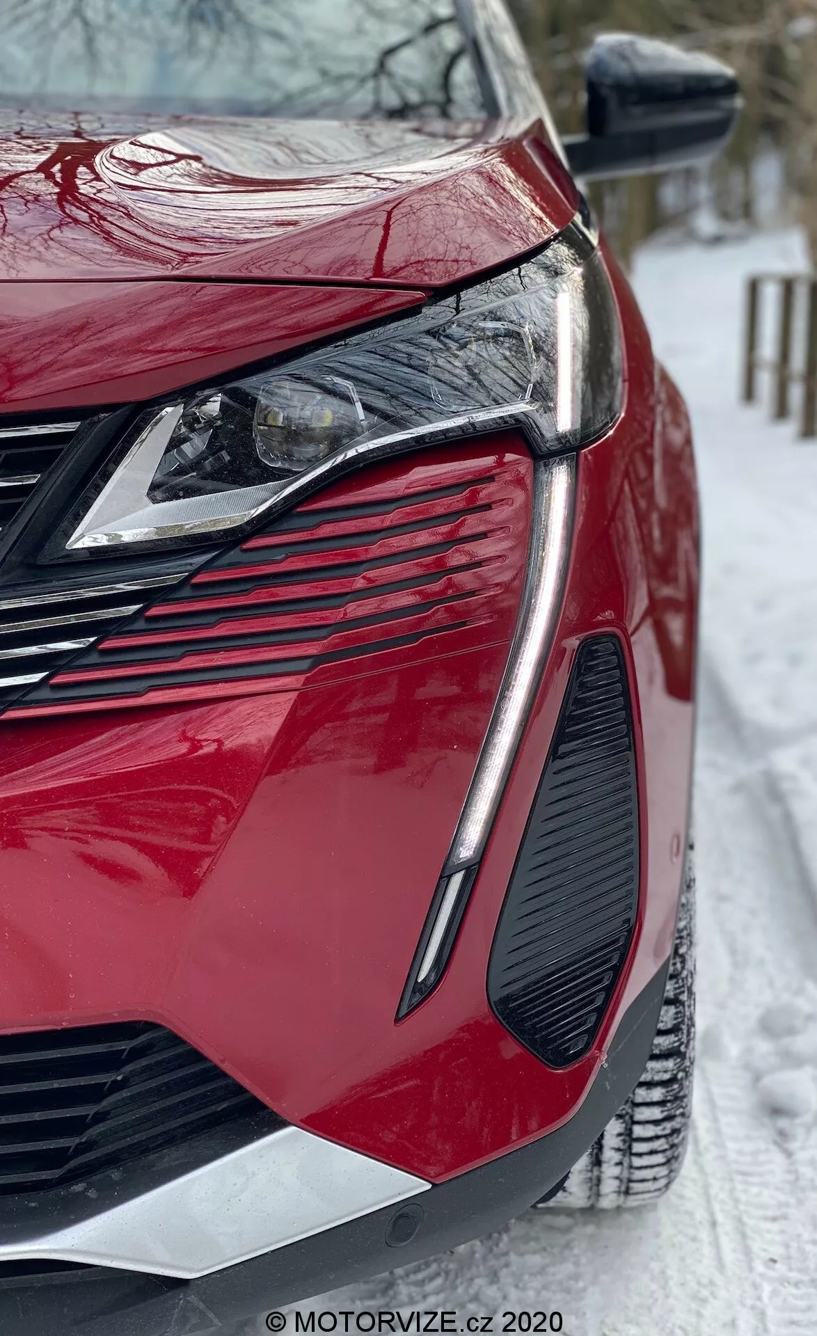 Detailný záber z nízkeho uhla na moderný červený Peugeot 3008 2021. Zamerané na prednú ľavú štvrtinu, ktorá ukazuje elegantné uhlové LED svetlomety, veľkú agresívnu mriežku, výrazný dizajn ventilačných otvorov, lesklý lak a ostro kontúrované panely karosérie. Bočné zrkadlo má integrované smerovky a scéna zahŕňa zasneženú zem, čo naznačuje zimné podmienky. Viditeľný vodotlač z MOTORVIZE.cz.