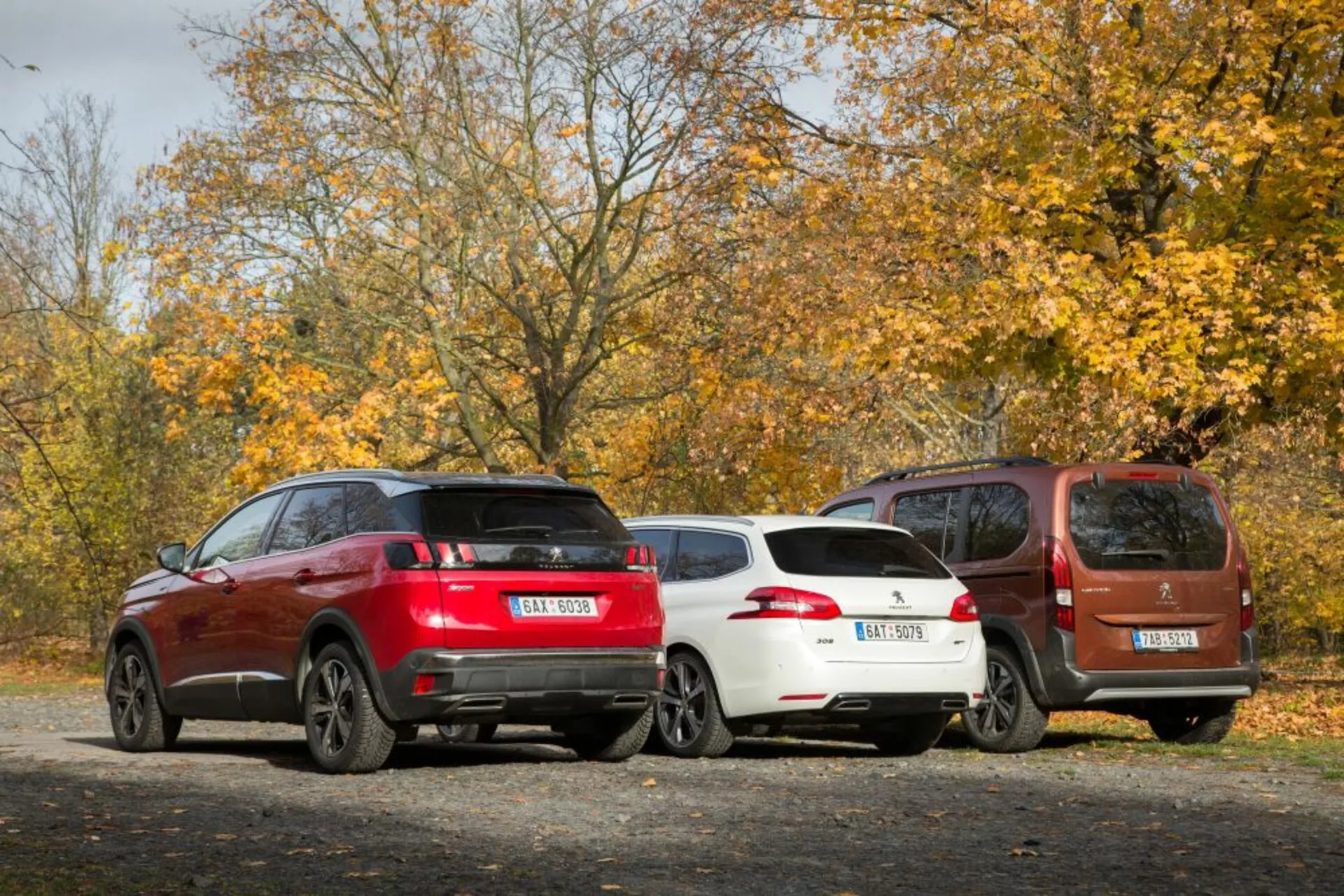 Zadný pohľad na tri zaparkované autá na štrkovej ploche s jesennou témou, vrátane červeného športového SUV, bieleho štýlového hatchbacku a hnedého kompaktného MPV, všetky vybavené modernými prvkami ako sú zliatinové kolesá a senzory parkovania vzadu, za prírodného denného svetla.