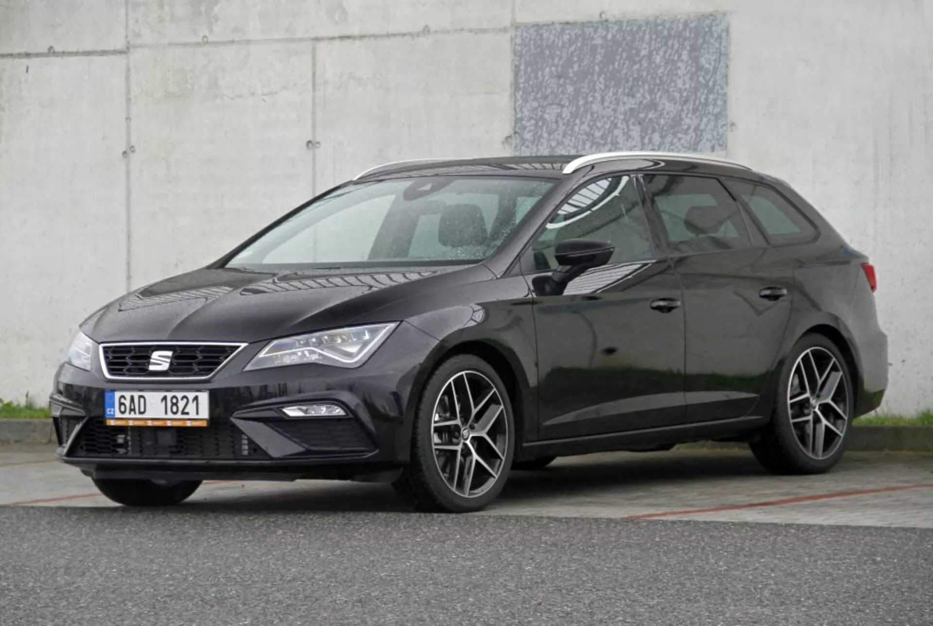 Hatchback Peugeot 308 SW tmavej farby z roku 2013 predvádzajúci svoje prvky: úzka mriežka s emblémom, elegantné svetlomety s dennými svetlami, športové kolesá z ľahkých zliatin s viacerými špicami, charakteristický bočný profil hatchbacku so zostupujúcou strechou, v kontrastnej tmavej farbe karosérie, odfotený z trojštvrťového predného uhla proti priemyselnému pozadiu