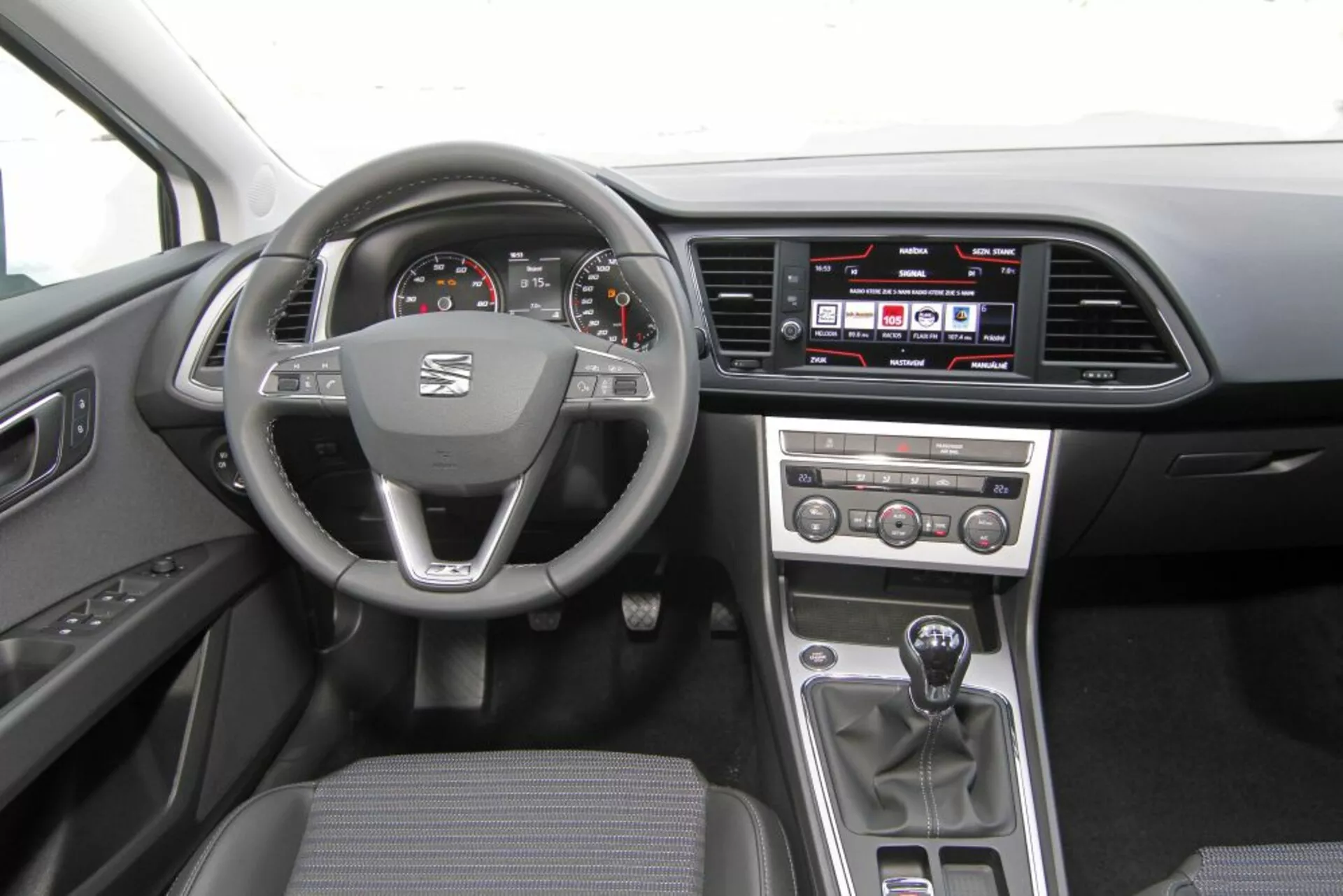 Interiér Peugeotu 308 SW z roku 2013 s volantom s logom a multifunkčnými ovládačmi, analógovým tachometrom a otáčkomerom s digitálnym displejom, dotykovým informačno-zábavným systémom na palubnej doske, tlačidlami pre manuálne ovládanie klímy na stredovej konzole, manuálnou radicou pákou a praktickým tmavým látkovým poťahom.
