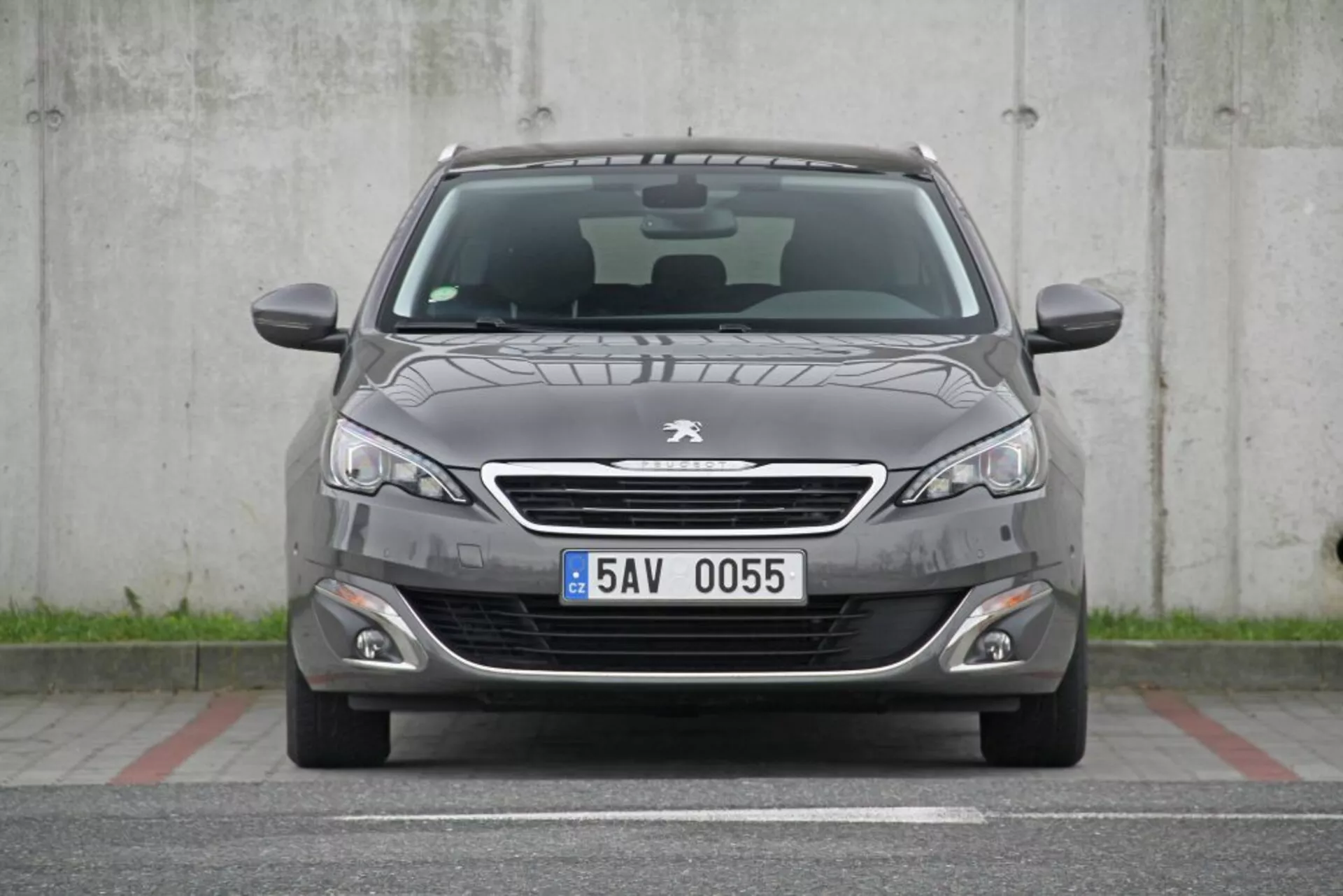 Čelný pohľad na Peugeot 308 SW ročník 2013 vo tmavosivej farbe s chrómovanou mriežkou, elegantnými prednými svetlami, hmlovými svetlami, športovým predným nárazníkom, jemnými záhybmi na kapote, viditeľnými stieračmi predného skla, európskou poznávacou značkou, parkovacími senzormi, včelou mriežkou na spodnej časti a viditeľnou panoramatickou strechou, zaparkovaný v betónovej oblasti bez prekážok