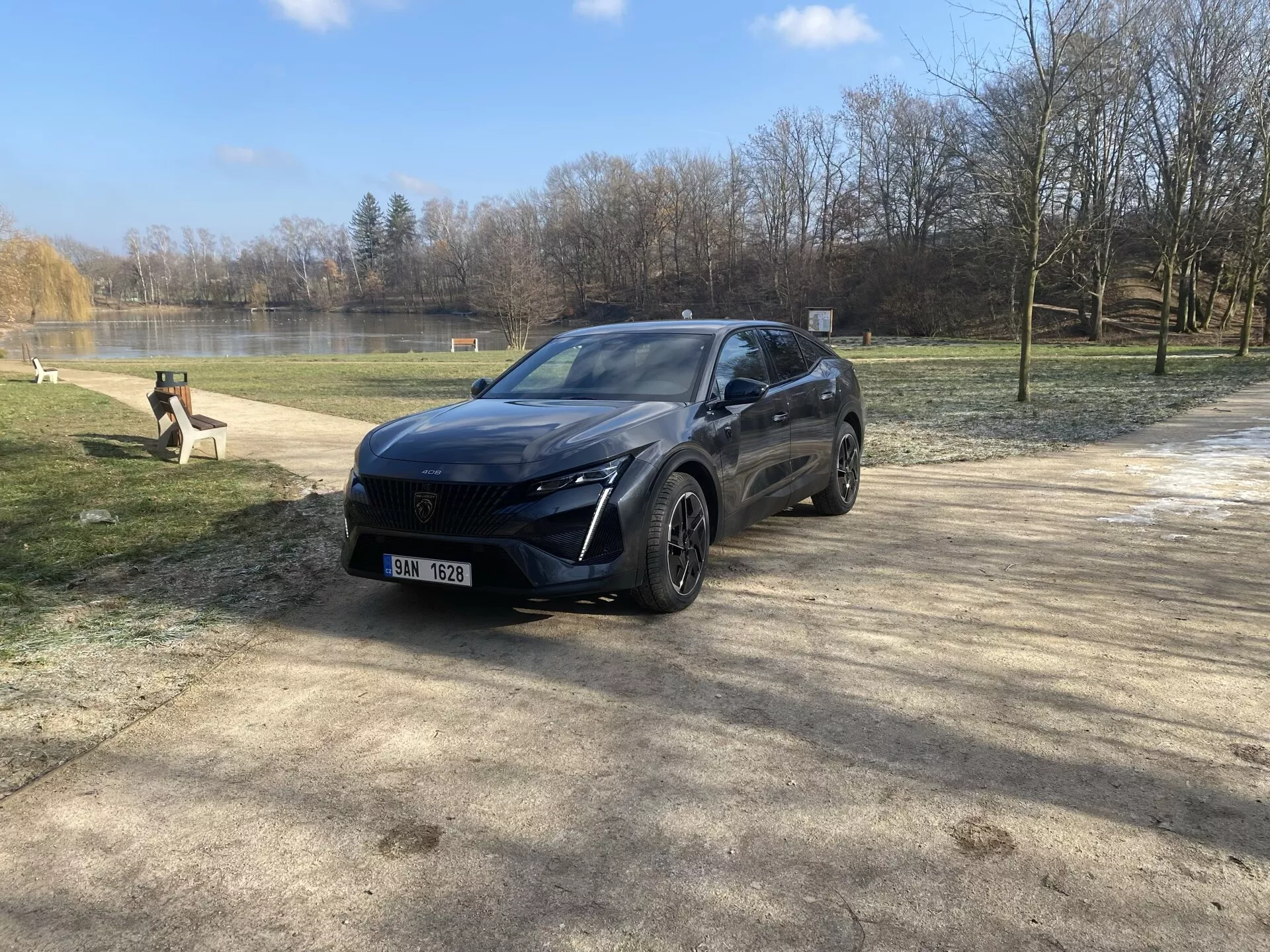 Peugeot 408 z roku 2023 zaparkované v prírodnom prostredí počas zimy alebo skorej jari, prezentujúce svoj moderný crossover dizajn, LED svetlomety, predný senzorový systém, zliatinové kolesá a prémiový finish z pohľadu z predného trojštvrťového uhla pod slnečným svetlom s odrazom šošovky.
