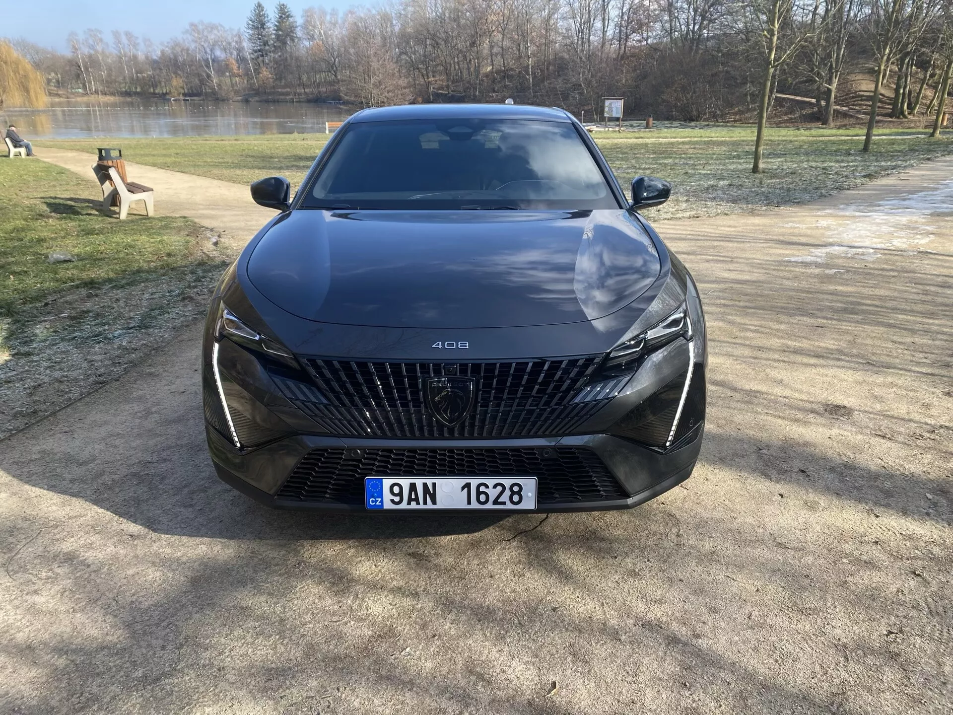 Predný pohľad na Peugeot 408 z roku 2023 zaparkovaný na štrkovej ploche s pozadím stromov a vody, zdôrazňujúci jeho veľkú mriežku s diamantovými vzormi, elegantné LED svetlomety, sochársky prepracované línie kapoty a pokročilé funkcie asistencie vodiča