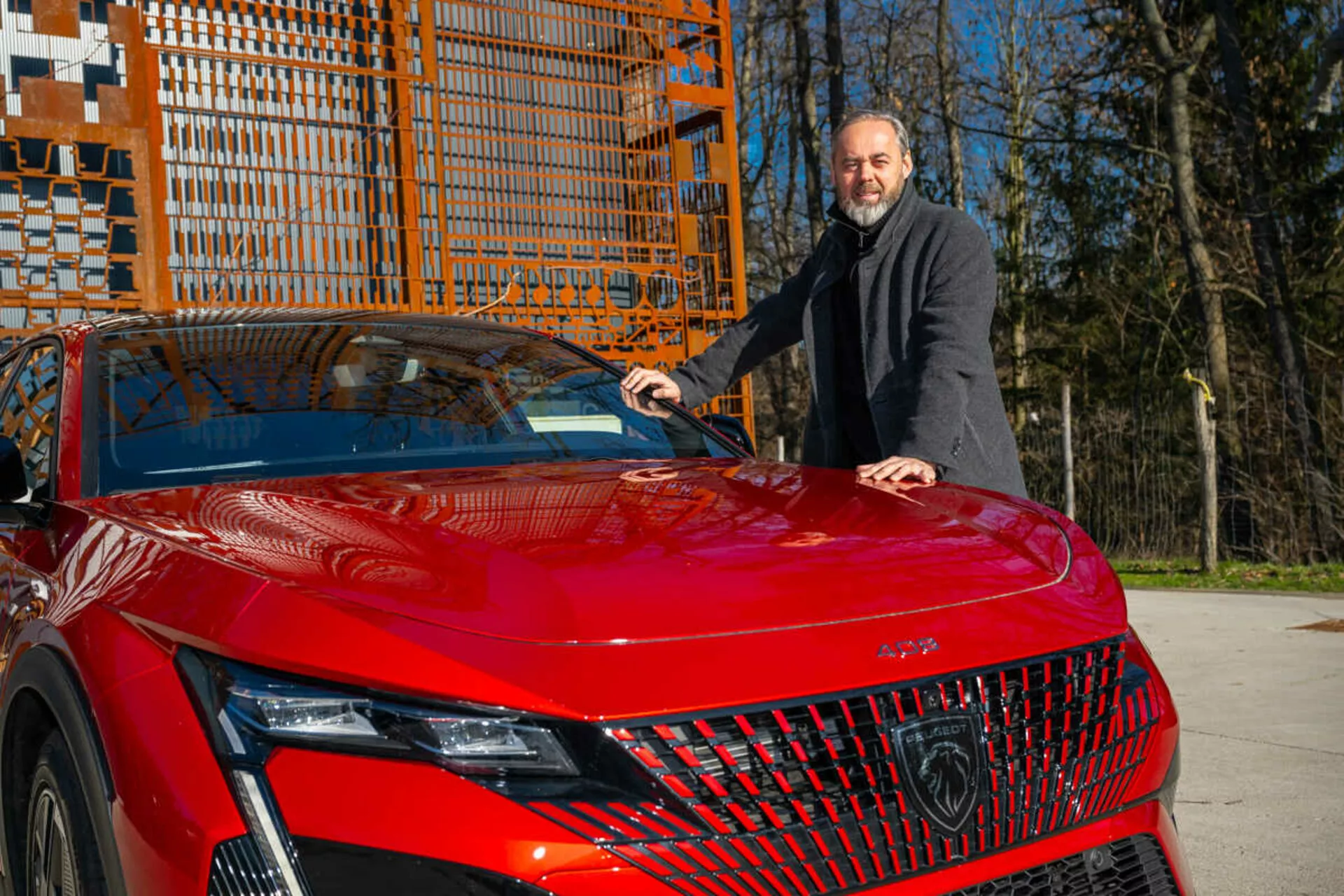 Červený model Peugeot 408 z roku 2023 predvádzajúci svoj predný ľavý štvrťrok na dennom svetle, zvýrazňujúci detailné LED svetlomety, geometrickú mriežku a športovo aerodynamickú kapotu s mužom v tmavom kabáte stojacim vedľa neho a kladúcim si ruku na kapotu