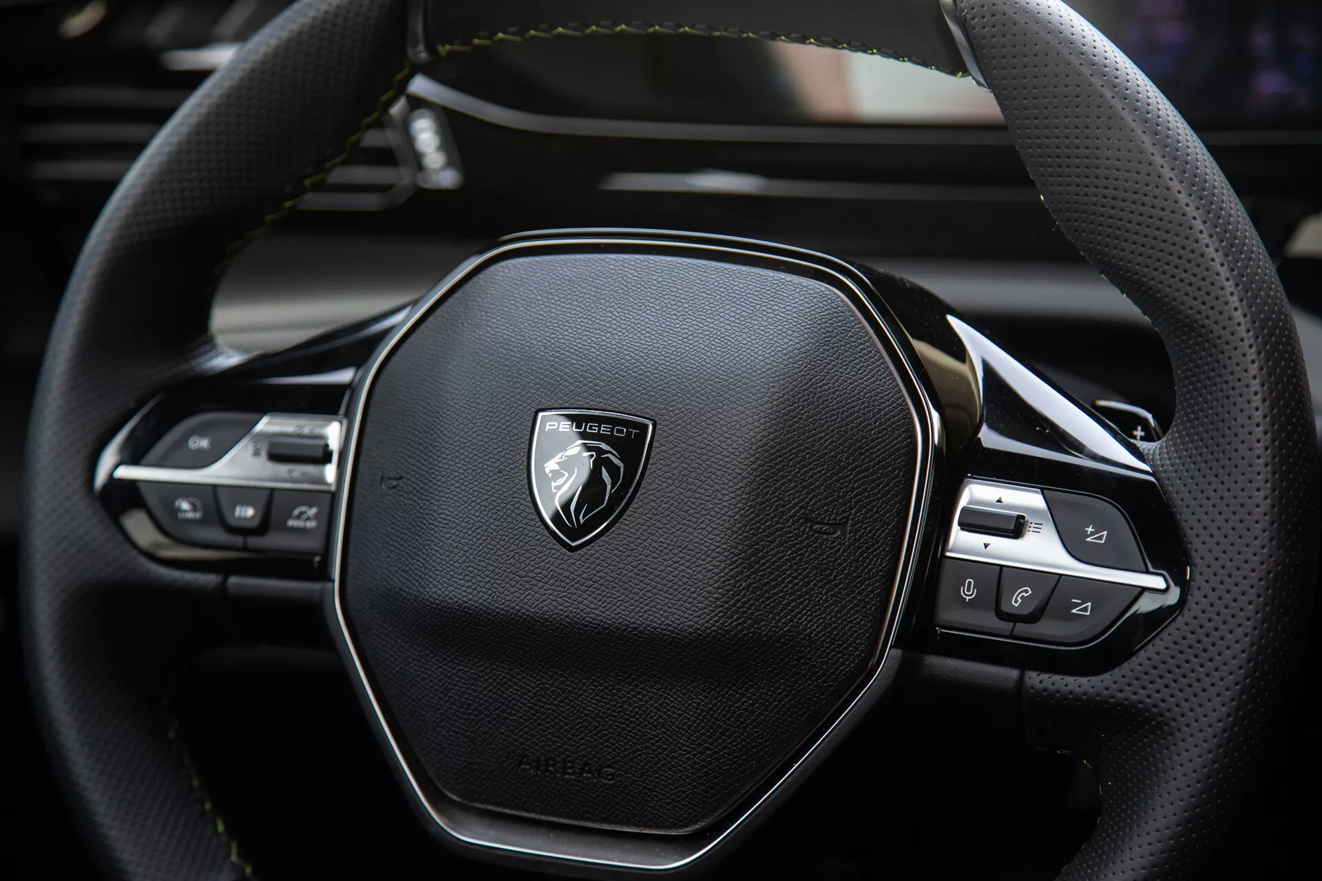 Detailný pohľad na volant Peugeot 408 2023 s ergonomickým šesťuholníkovým dizajnom, prešívaním z kože, integrovanými ovládacími prvkami pre audio a nastavenia tempomatu, s emblémom na brúsenej kovovej doske.