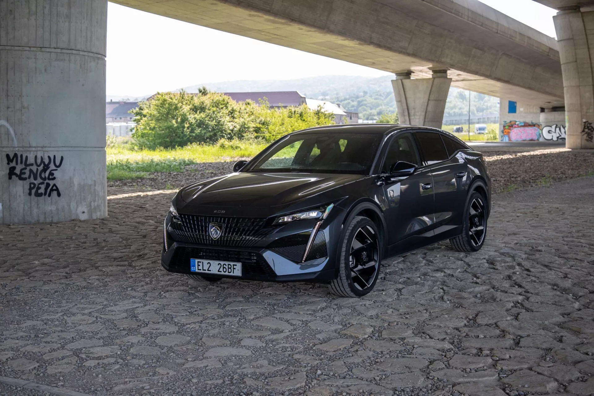 Peugeot 408 2023 zachytený z uhla tri štvrtiny zdôrazňujúci jeho hladké LED svetlomety, vzorovanú mriežku a športový aerodynamický predný nárazník pod prírodným denným svetlom na spevnenej ploche pod nadjazdom