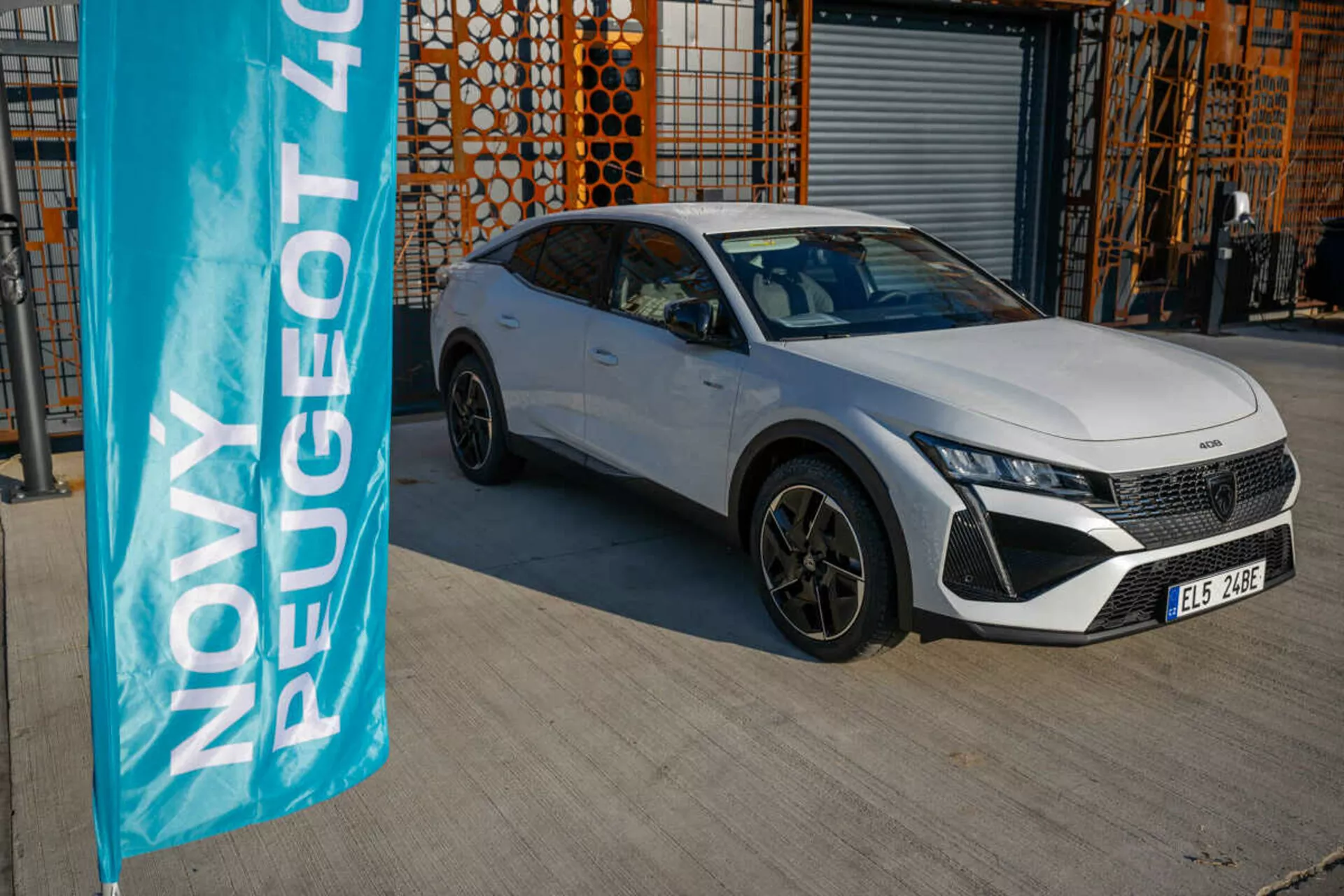 Biely Peugeot 408 ročník 2023 zaparkovaný vonku s priemyselnou architektúrou na pozadí, predvádzajúci dizajnové a technologické funkcie vozidla pod prirodzeným denným svetlom
