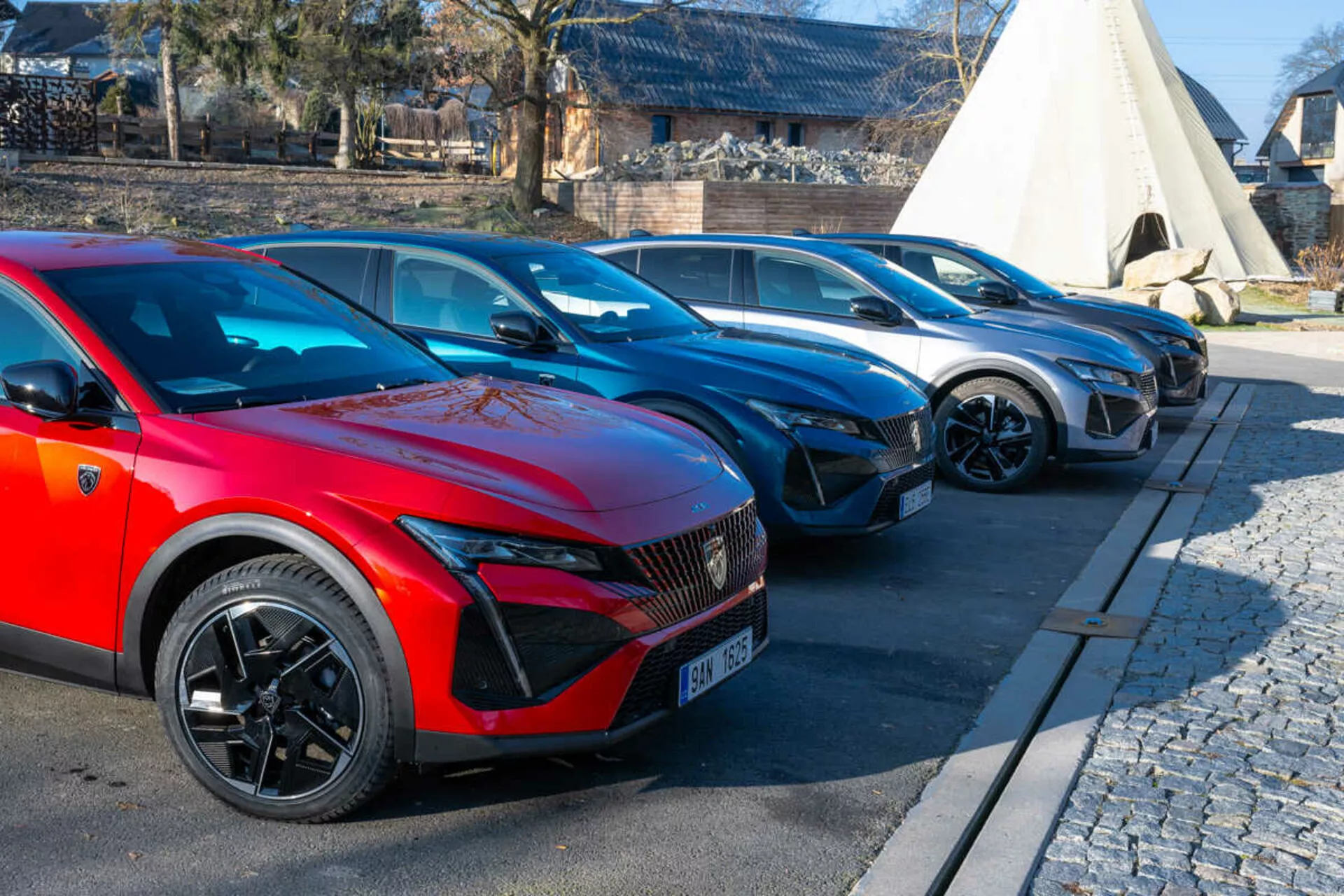 Rad vozidiel Peugeot 408 z roku 2023 s červeným autom v popredí so športovými viacramennými kolesami a LED svetlometmi naznačujúcimi elektrický alebo hybridný model, zaparkovaný na vydláždenej ploche vedľa dlažby