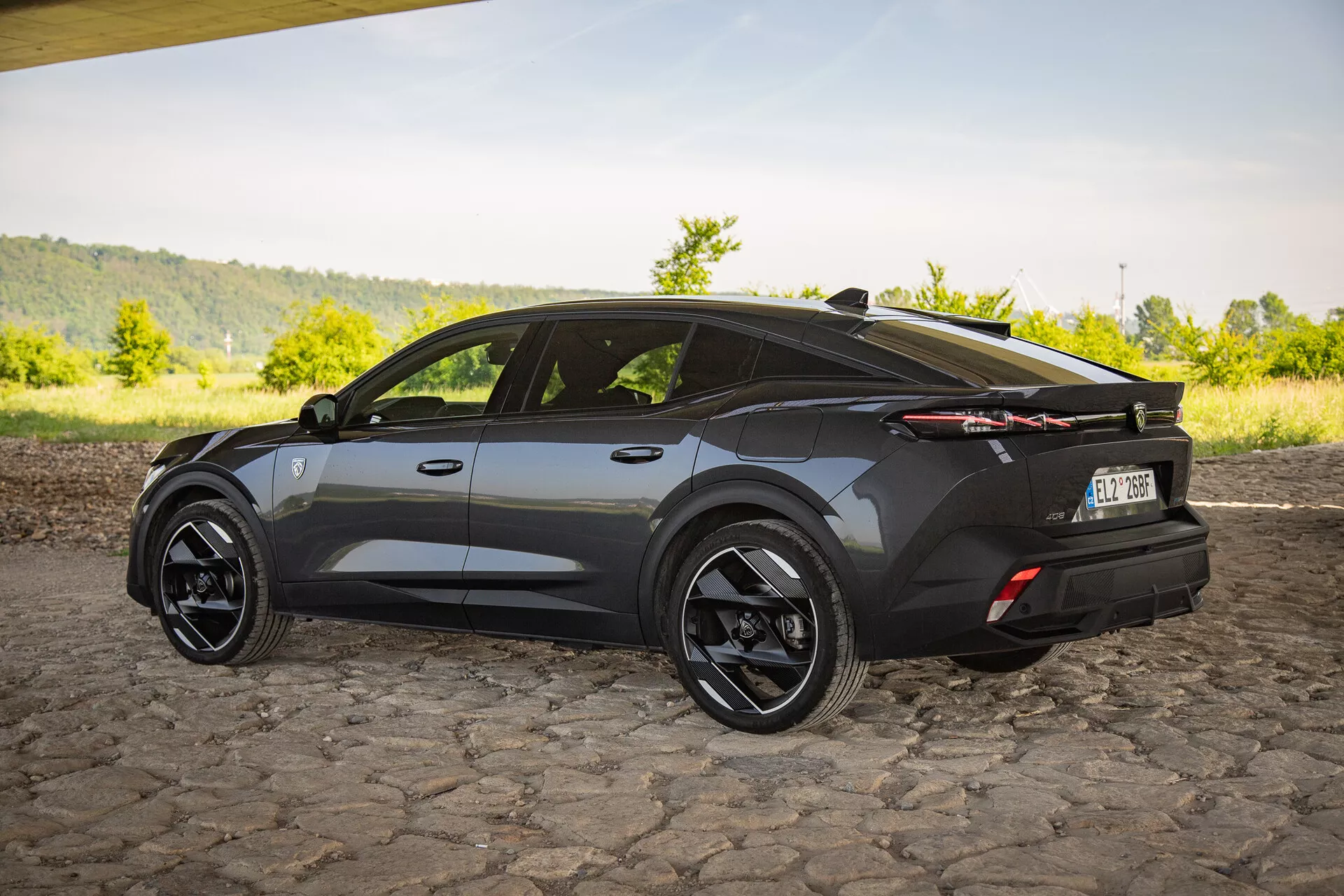 Crossover SUV Peugeot 408 z roku 2023 vo tmavom prevedení, s trojštvrtečným pohľadom zozadu zdôrazňujúcim aerodynamický dizajn s ostrými uhlami, športový spádový tvar strechy, veľké čierne zliatinové kolesá, výrazné LED zadné svetlá a decentné zadné krídlo na dlažobnom povrchu