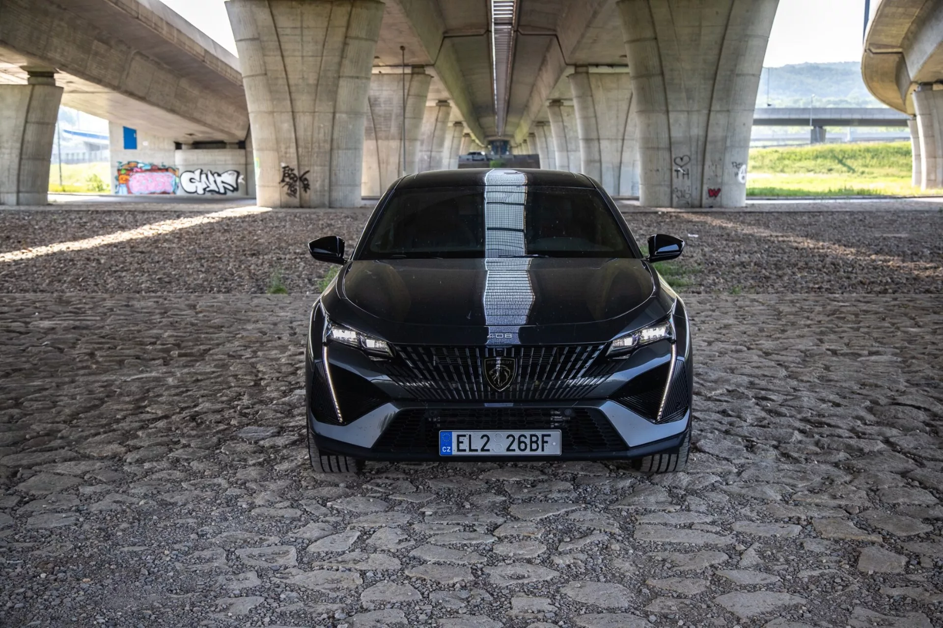 Peugeot 408 z roku 2023 v dynamickom pohľade spredu zdôrazňujúcom jeho agresívny postoj a športový dizajn s detailnou mriežkou, štíhlymi LED svetlometmi a aerodynamickými prvkami na vzorovanej dlažbe pod mestským mostom