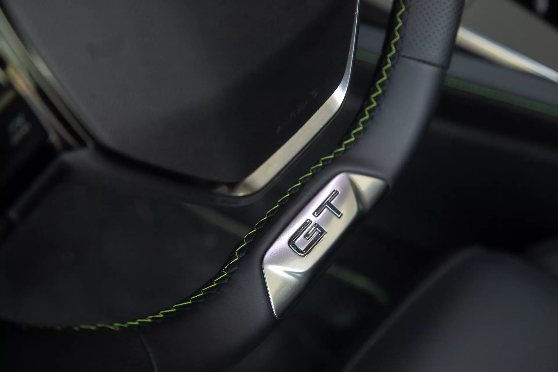 Detailný pohľad na časť volantu a palubnej dosky Peugeot 408 2023, čierny kožený volant so zeleným prešívaním a znakom GT, zdôrazňuje luxus a športovosť