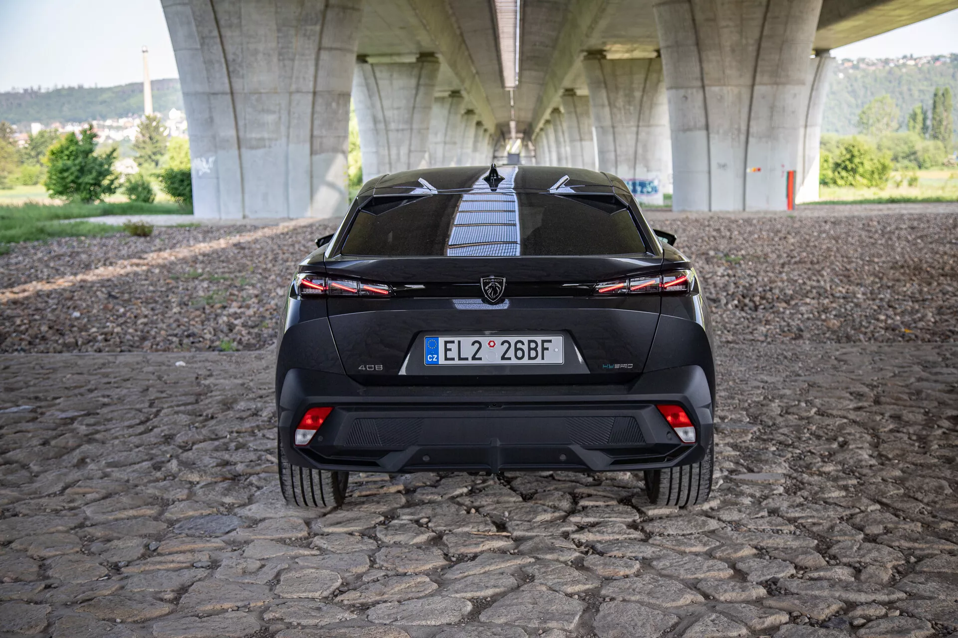 Zadný pohľad na Peugeot 408 2023 ukazujúci zadné dvere, nárazník, spodný difúzor, zadné svetlá so spojovacou svetelnou lištou a senzory na pomoc pri parkovaní, fotografované pod mostom na povrchu z kockovej dlažby