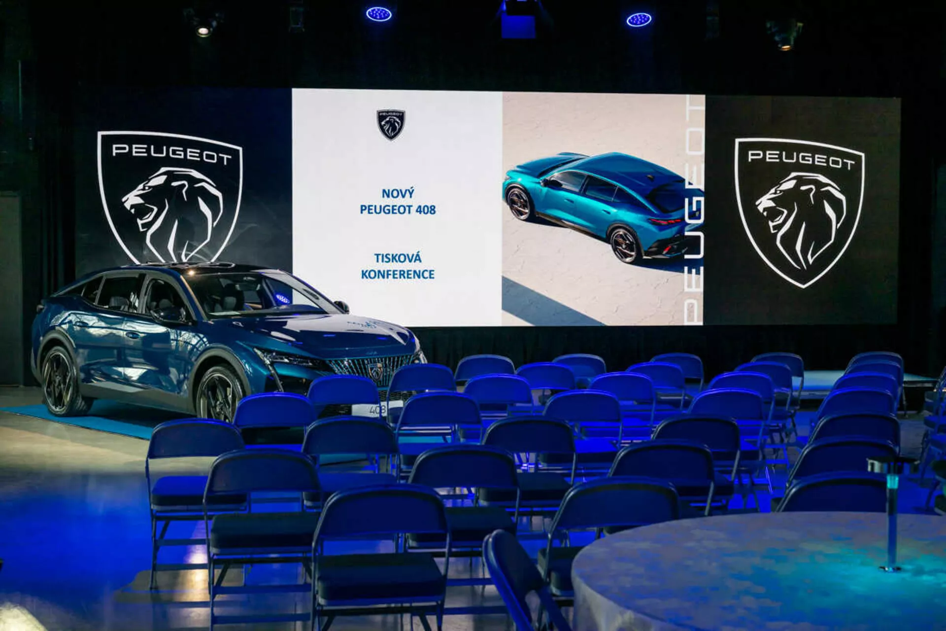 Peugeot 408 prezentované na tlačovej konferencii, zobrazené pod 45-stupňovým uhlom zdôrazňujúce jeho predné a bočné profily, úhľadný dizajn a LED svetlomety, s digitálnymi obrazovkami a značením na pozadí
