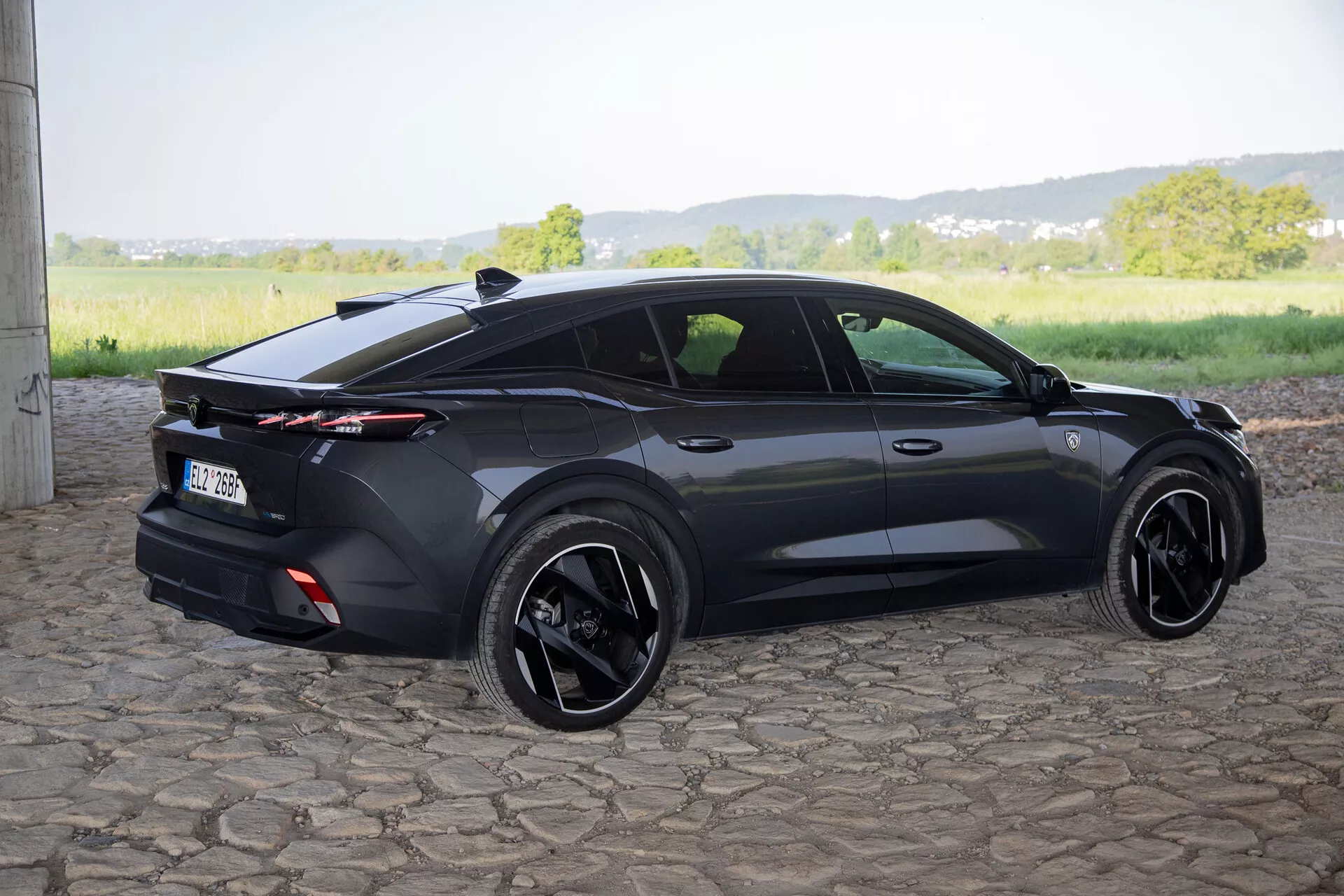 Peugeot 408 z roku 2023 zachytený z uhla tri štvrtiny zozadu, čo zdôrazňuje jeho športový dizajn hatchbacku, integrovaný zadný spojler s celoplošnými zadnými svetlami, čierne zliatinové kolesá a súčasné prvky ako sú zapustené kľučky dverí a bočné zrkadlá so smerovkami, na prírodnom dennom svetle