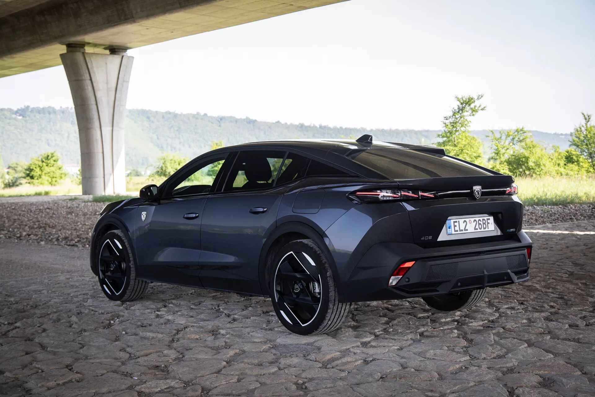 Trojštvrťový zadný pohľad na hatchback Peugeot 408 rok 2023 pod mostom na dlažobných kockách so športovými diskami a pokročilou svetelnou technológiou