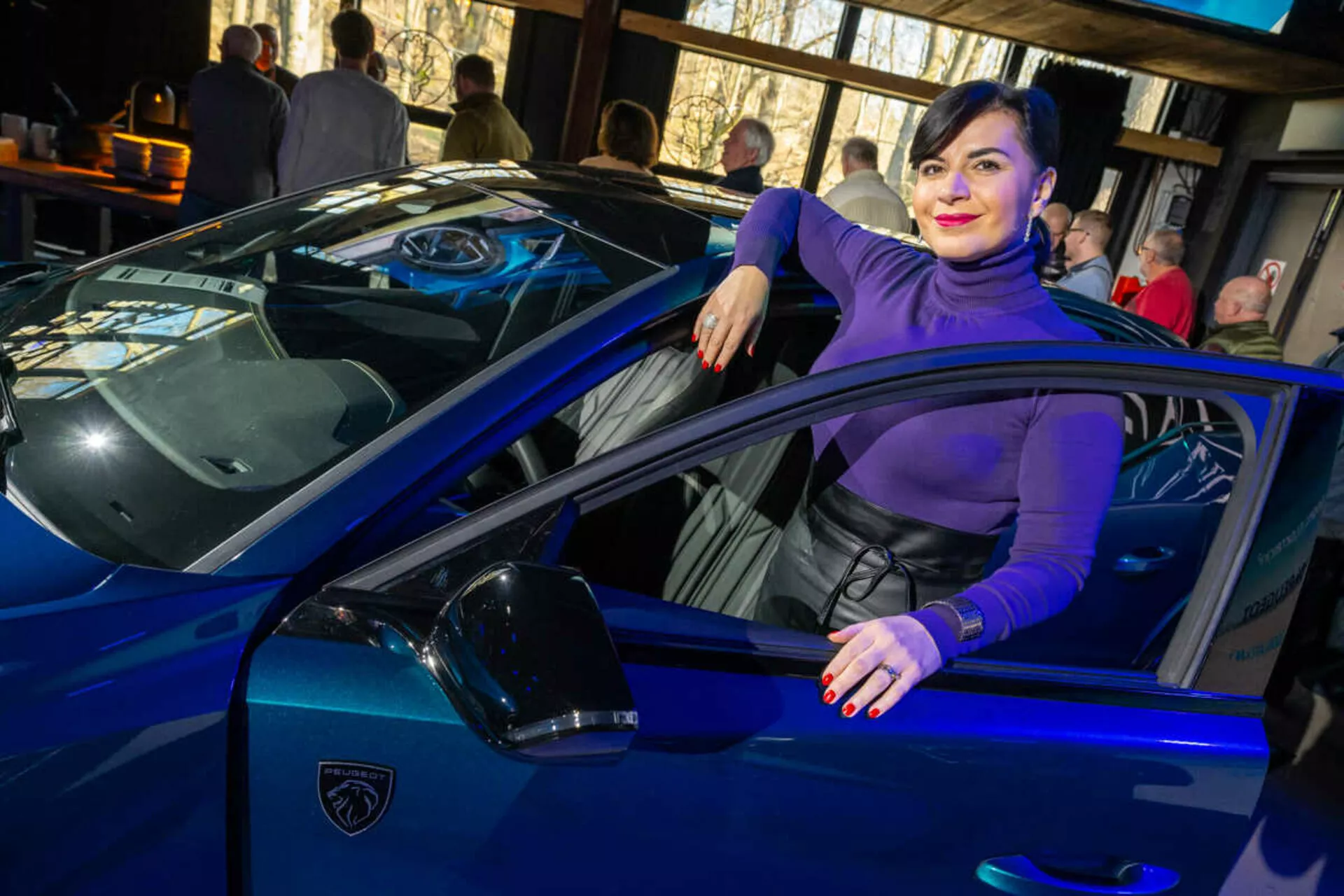 Živé modré športové auto, pravdepodobne Lamborghini, zachytené z uhla tri štvrtiny zdôrazňujúce jeho štíhle línie, ostré LED svetlomety a ikonické logo v tvare štítu, s osobou neformálne interagujúcou s vozidlom naznačujúcou luxus a štýl.
