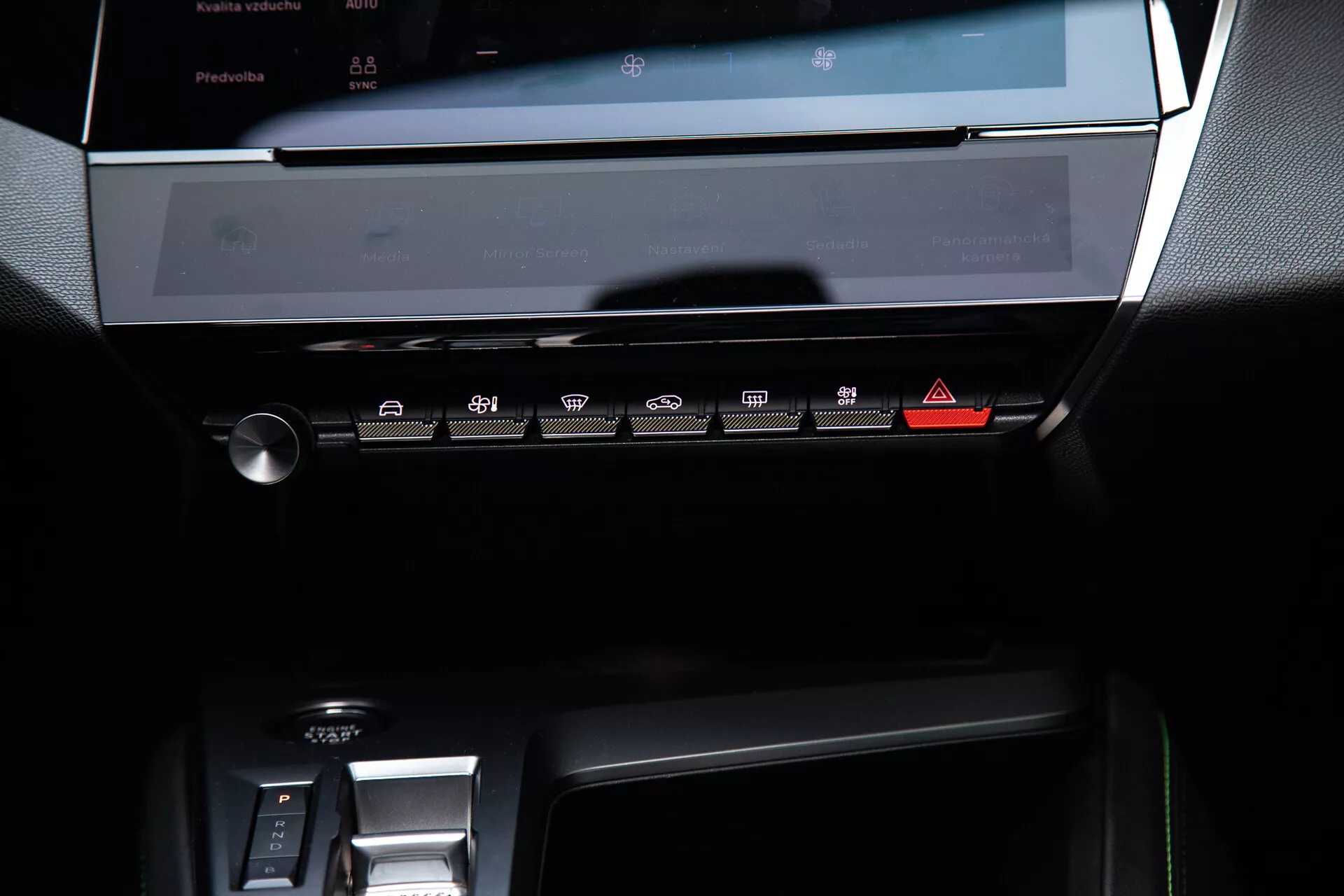 Centrálna palubná doska Peugeotu 408 z roku 2023 so vypnutým dotykovým displejom infotainment systému, panelom klimatizácie s dotykovými tlačidlami a časťou oblasti elektronického radenia, pod jemným ambientným osvetlením zdôrazňujúcim jej moderný, elegantný dizajn s tmavými tónmi a kovovými akcentmi.