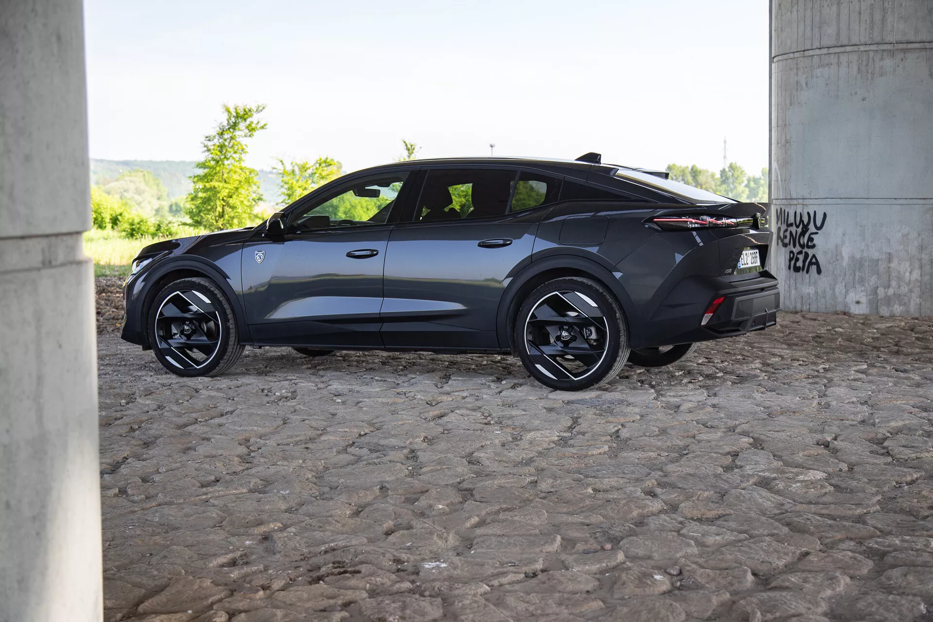 Profilový pohľad na moderné tmavé auto Peugeot 408 2023 pod mostom s graffiti, vynikajúce aerodynamickým dizajnom, zliatinovými kolesami a tónovanými oknami