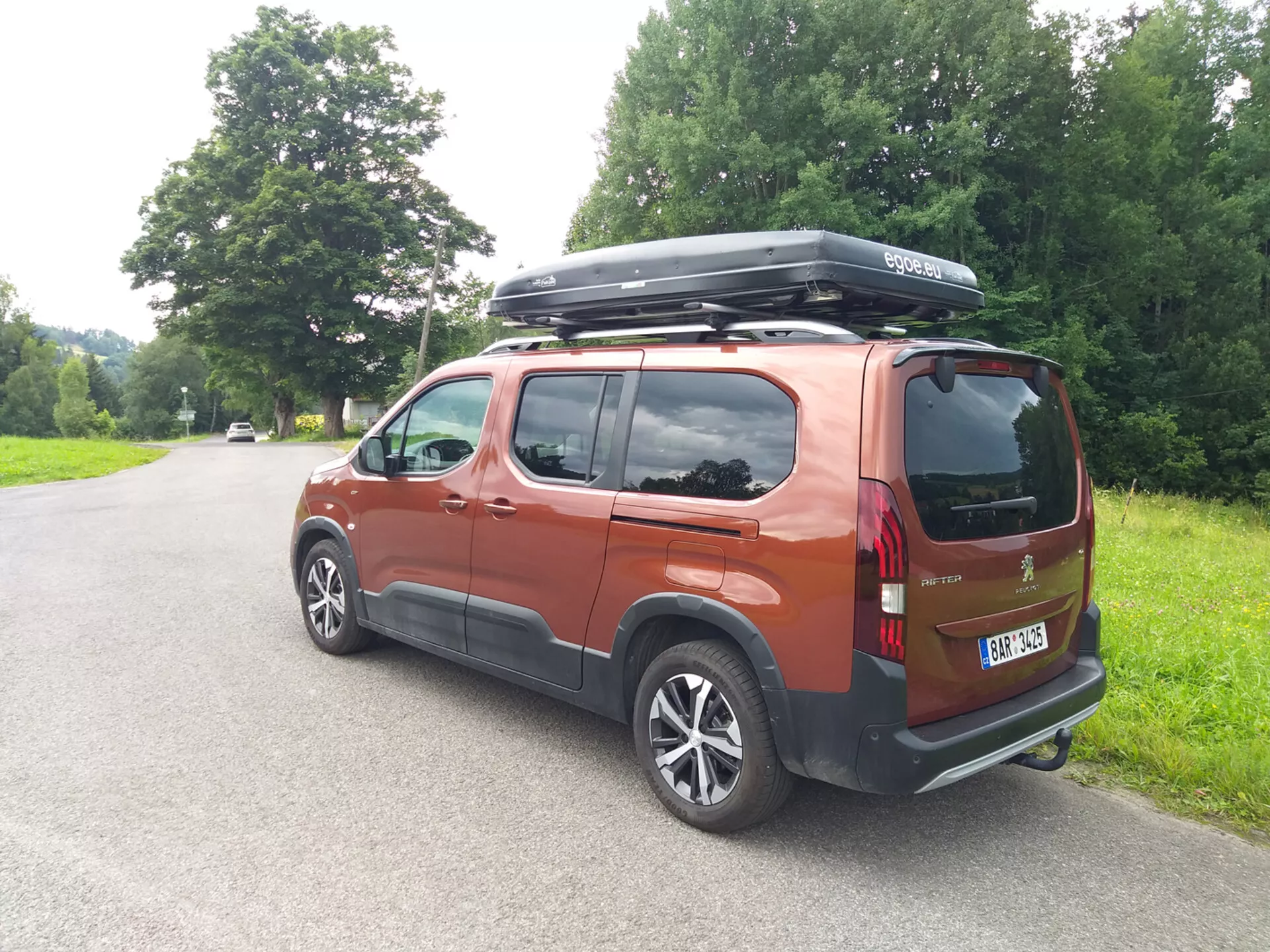 Zadný trojštvrťový pohľad na Peugeot Rifter 2018 v metalickej páleno-oranžovej farbe, zaparkovaný na vidieckej ceste, s hliníkovými kolesami, LED zadnými svetlami, strešným boxom, sklami pre súkromie a krátkou anténou za denného prírodného osvetlenia