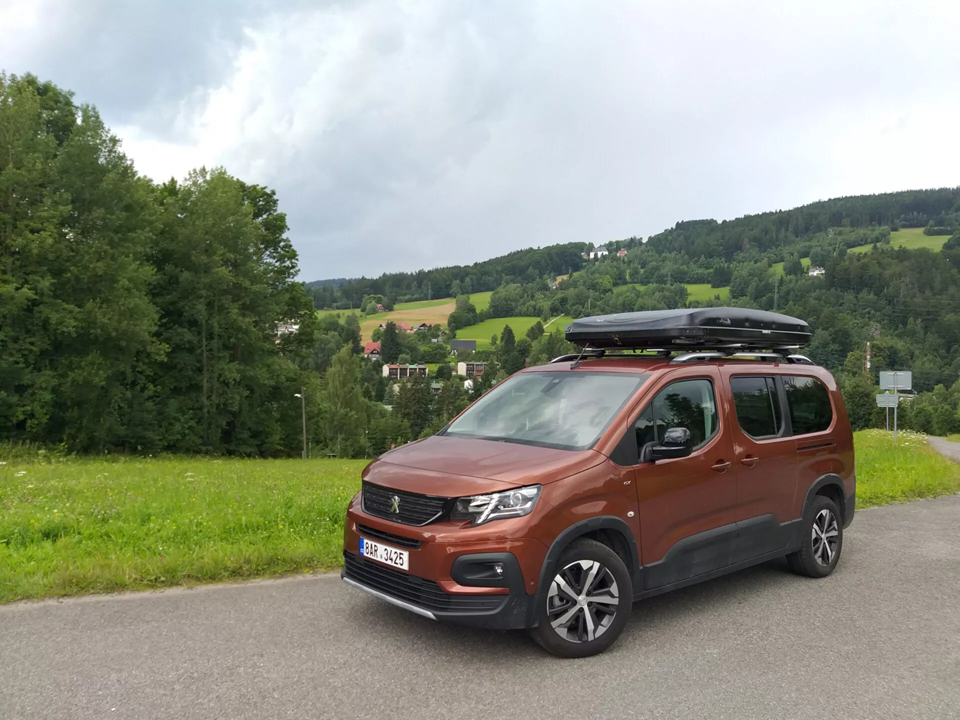 Bočný predný pohľad na Peugeot Rifter 2018 v metalickej bronzovej farbe, ukazujúci výraznú mriežku chladiča, súčasné LED predné svetlá, hmlové svetlá, zliatinové kolesá, vonkajšie spätné zrkadlá v farbe karosérie, bočné okná s potenciálnym súkromným sklom, systém strešných nosičov, priestranný interiérny profil, plastovú ochranu karosérie pre mierne off-road použitie a bočné posuvné dvere, zaparkovaný na asfaltovej ceste so zeleňou a kopcom na pozadí, za podmienok zamračeného osvetlenia.