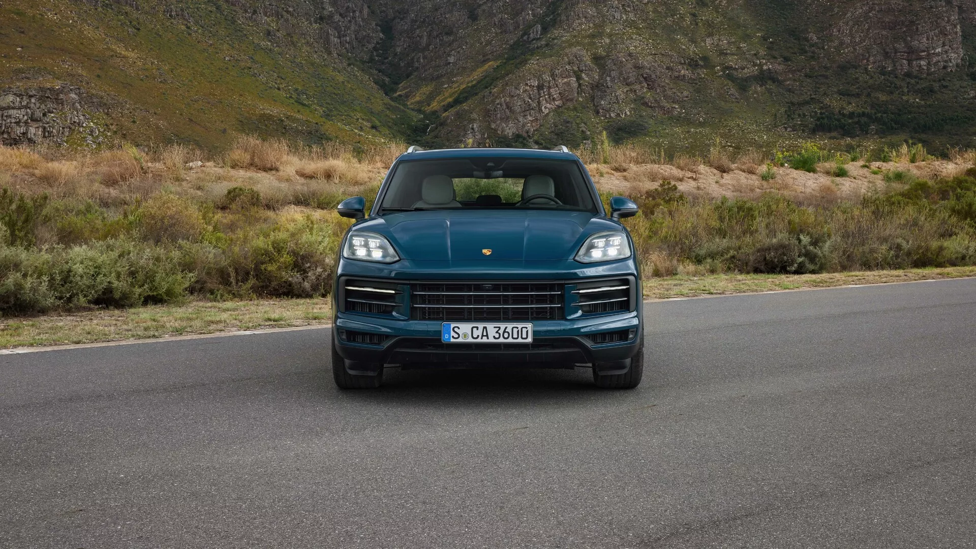 Porsche Cayenne 2023 (9YA Facelift) z pohľadu priamo z očí ukazuje LED svetlomety, výraznú masku s logom značky, športový dizajn kapoty a integrované parkovacie senzory na pozadí horskej krajiny, čo naznačuje luxus a dobrodružstvo