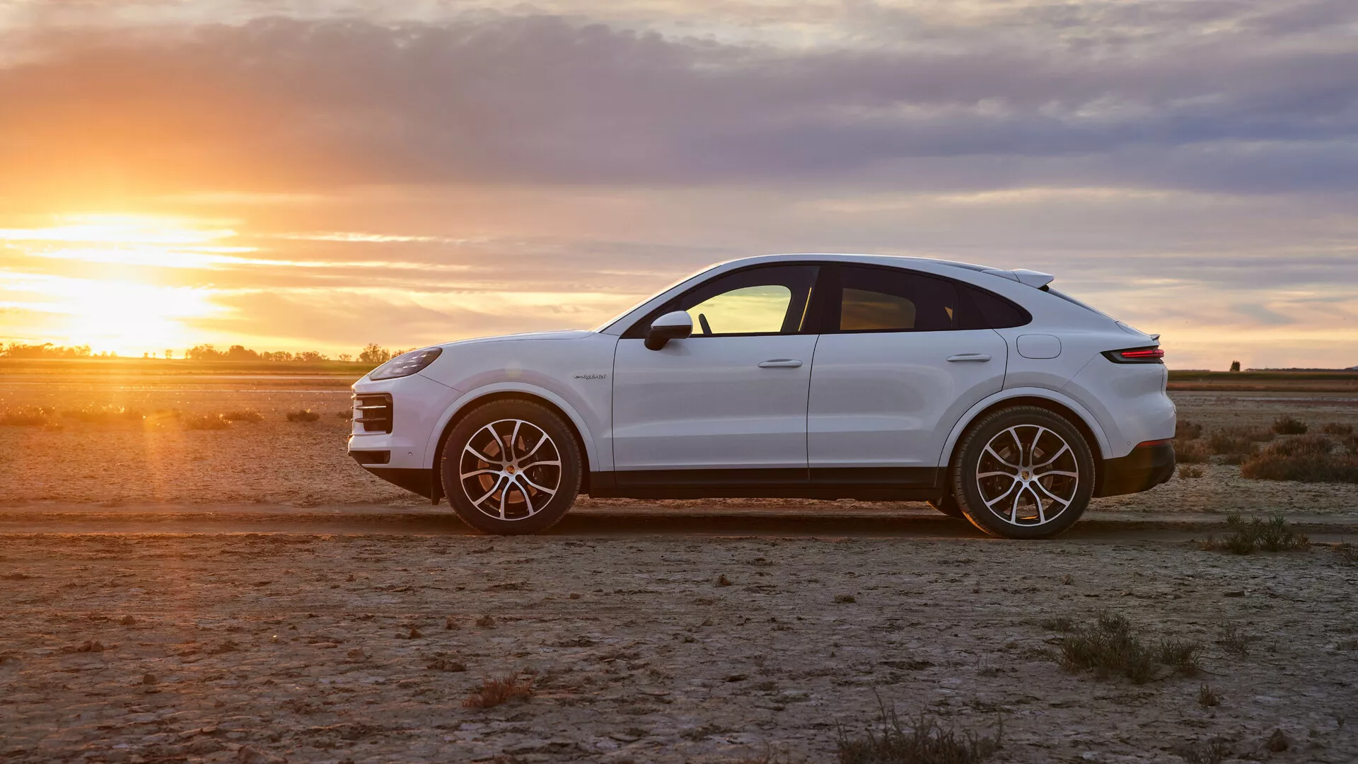 Luxusné crossover SUV Porsche Cayenne 2023 v lesklej bielej farbe na aridnom teréne počas zlatej hodinky, zobrazujúce profilový pohľad, športové zliatinové kolesá a štýlové dizajnové prvky
