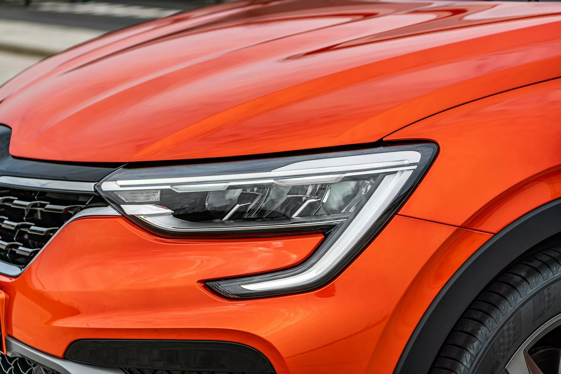 Detailný záber na prednú časť jasne oranžového vozidla Renault Arkana 2021, zameraný na moderné dizajnové detaily, vrátane pokročilých LED svetlometov, aerodynamické krivky kapoty, čiastočne viditeľnú tmavú mriežku v tvare plásta medu a fragment veľkého zliatinového kolesa, naznačujúci prémiový výkon a špičkovú automobilovú technológiu.