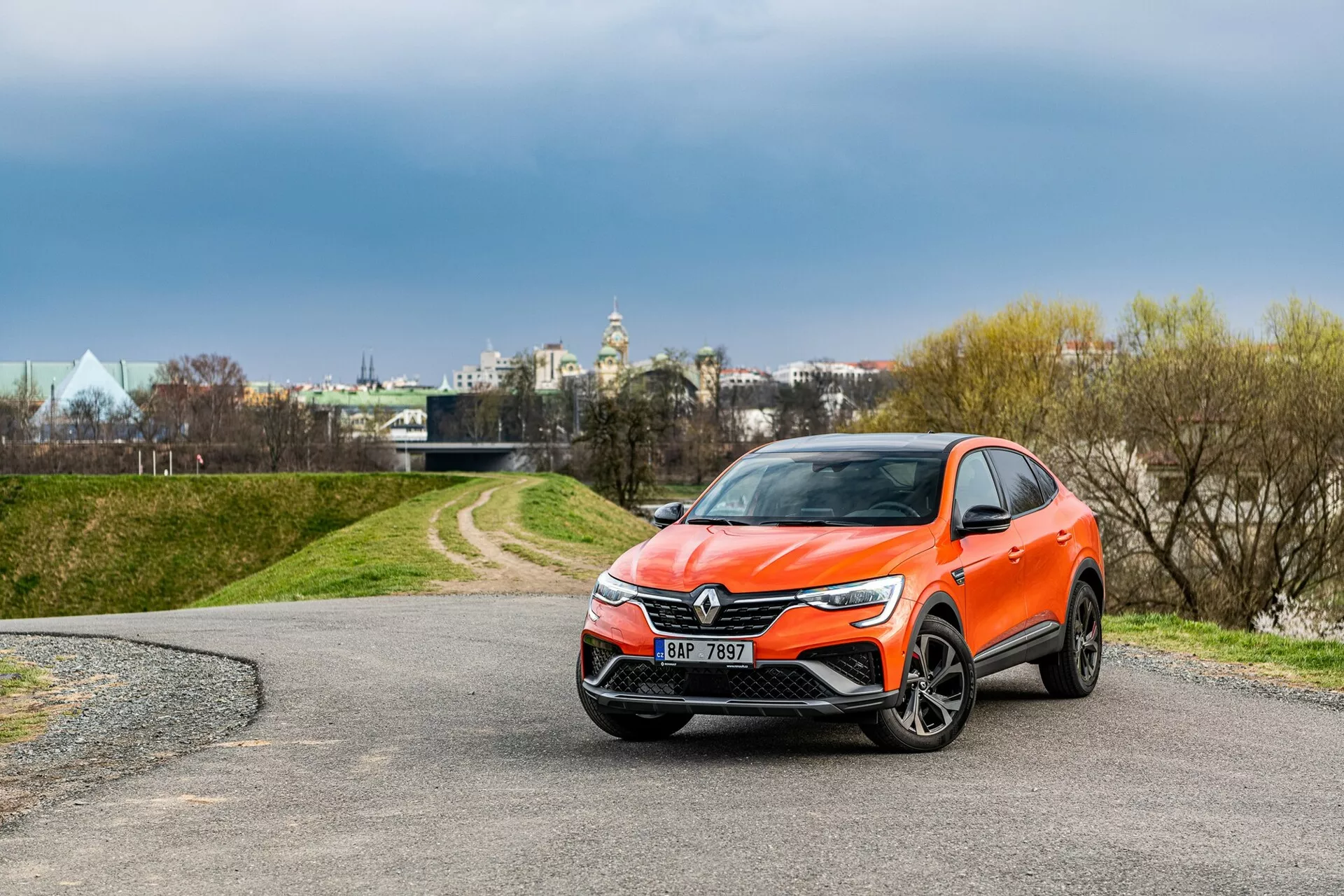 Obrázok zobrazuje moderné crossover SUV Renault Arkana 2021 zaparkované na kľukatej ceste. Snímka z trojštvrťového predného pohľadu jasne ukazuje prednú mriežku s výrazným emblémom, elegantné svetlomety siahajúce k stranám a presne štýlizovaný predný nárazník pravdepodobne vybavený hmlovými svetlami a senzormi. Aerodynamicky navrhnuté bočné zrkadlá s možnými integrovanými smerovými svetlami, zliatinové kolesá a jasná oranžová farba karosérie zvyšujú celkový vzhľad. Prostredie zahŕňa stromy a zamračenú oblohu v pozadí, so štruktúrami naznačujúcimi mestskú oblasť v diaľke.