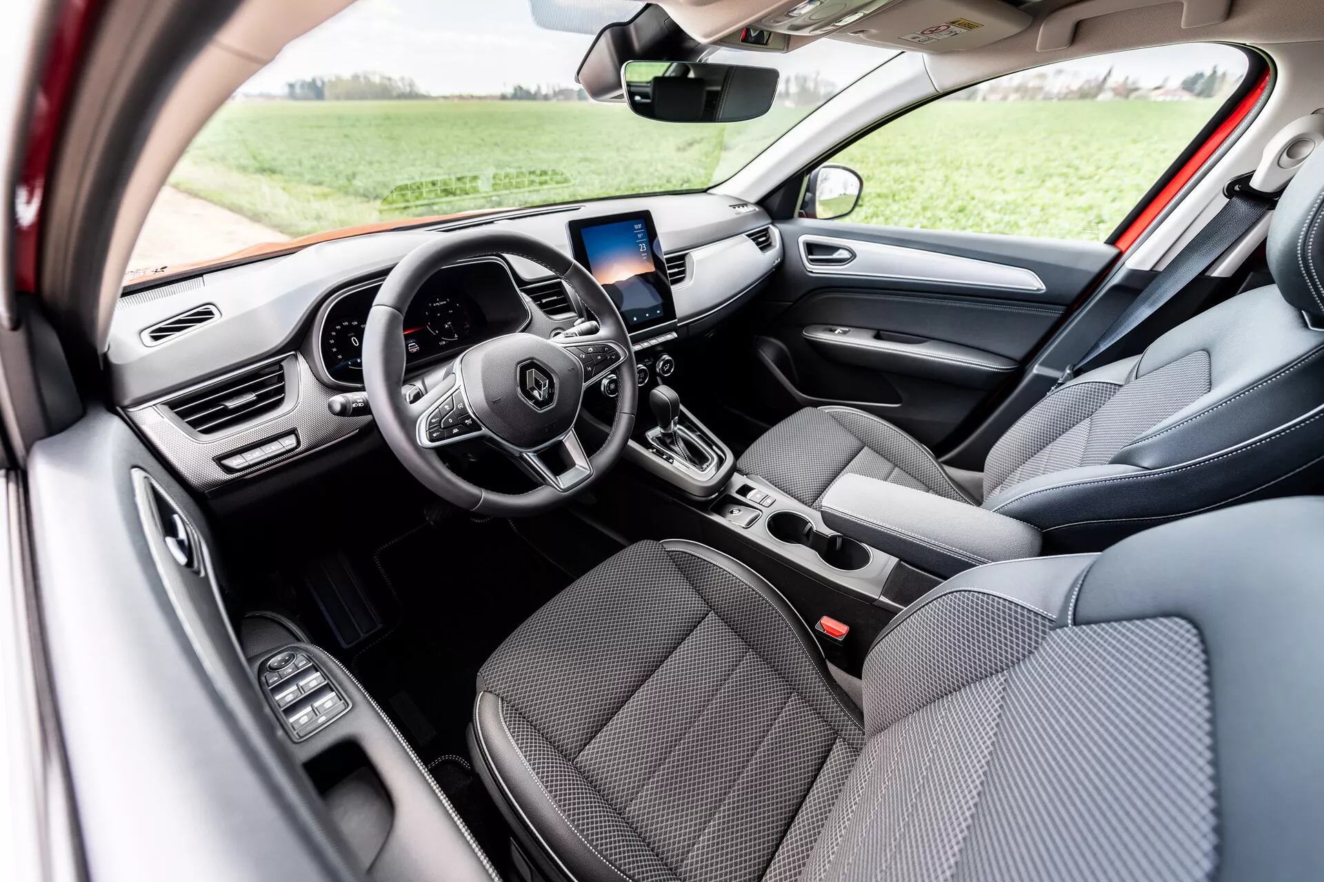 Interiér Renault Arkana 2021 so sedadlami potiahnutými sivou látkou vpredu, multifunkčným volantom s tromi ramenami s integrovanými ovládacími tlačidlami, digitálnym displejom palubnej dosky, obrazovkou infotainmentu s navigáciou GPS nad centrálnymi ventilmi, manuálnymi ovládacími prvkami klimatizácie, automatickou radenou pákou a ovládaním elektrických okien na dverách vodiča, osvetlené prirodzeným denným svetlom v exteriérovom prostredí