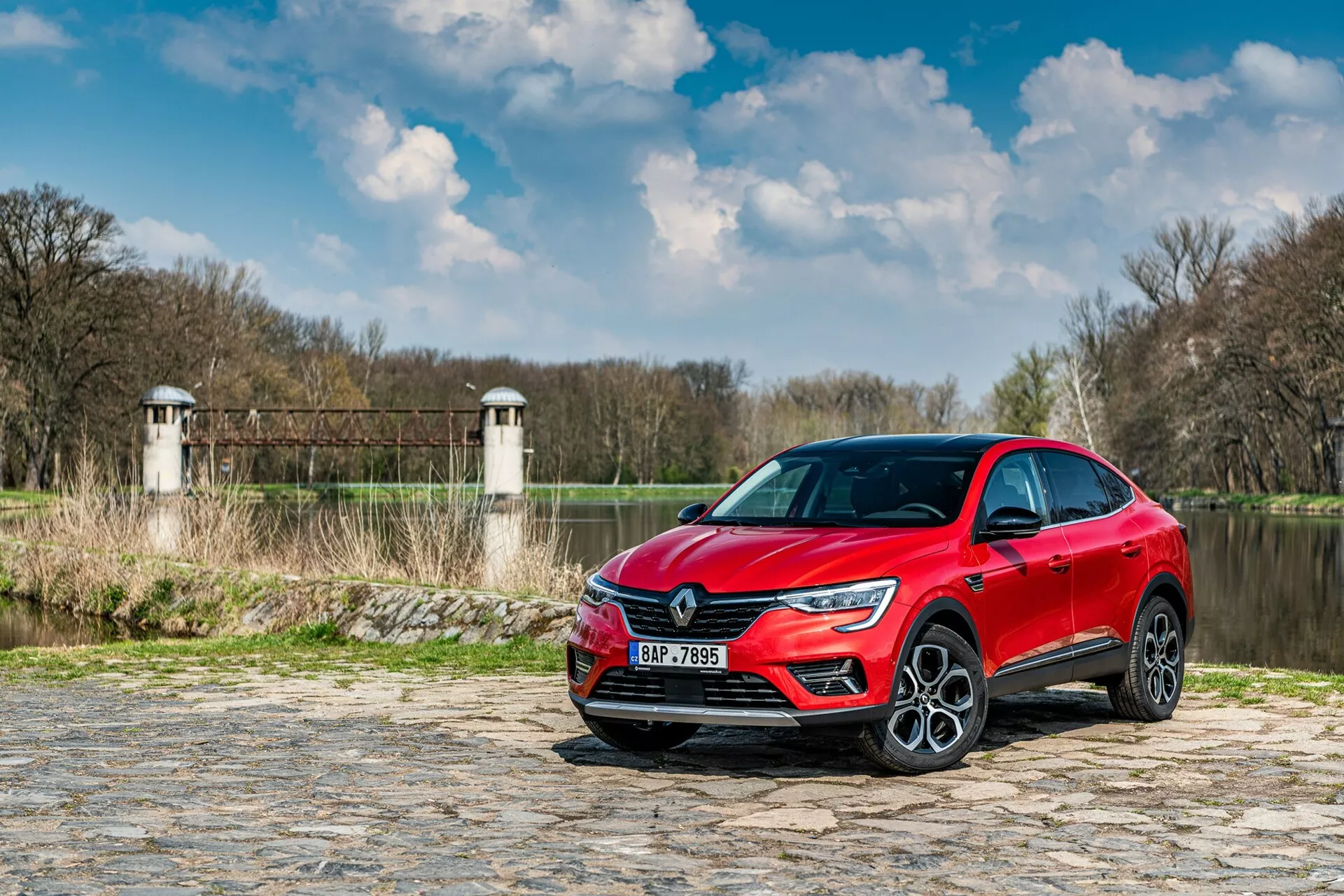 Obrázok zobrazuje Renault Arkana 2021 zaparkovaný na dláždenom chodníku pri rieke alebo jazere, s vegetáciou a štruktúrami podobnými vežiam hrádze alebo mostného systému v pozadí. Auto je zachytené z nízkeho uhla, trištvrťový predný pohľad, zvýrazňujúc prednú masku, LED svetlomety a časť bočného profilu vrátane dverí vodiča a predného blatníka. Moderné dizajnové prvky, ako ostrý svetelný podpis LED a veľké zliatinové kolesá, naznačujú vyššiu úroveň výbavy. Reflexná metalická farba pridáva estetický pôvab, zatiaľ čo možné senzory asistenta vodiča a technológia zabudovaná v nárazníkoch a bočných zrkadlách zlepšujú funkčnosť.