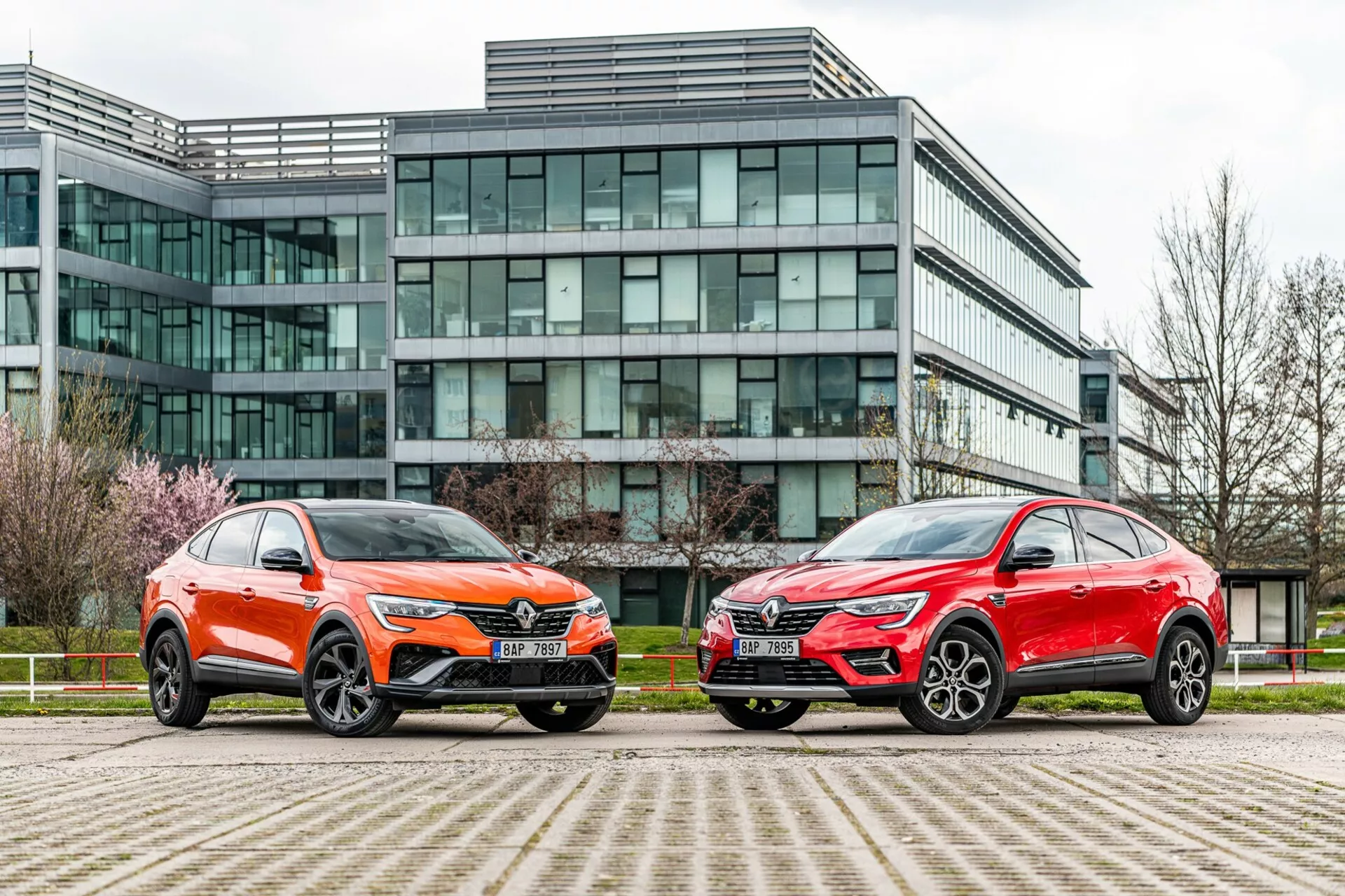 Obrázok zobrazuje dve SUV vozidlá vedľa seba zaparkované pred modernou kancelárskou budovou so sklenenou fasádou. Vozidlo vľavo je vo výraznej oranžovej farbe s kovovým leskom a má charakteristickú prednú mriežku, ostré LED svetlomety, tvarovanú kapotu a veľké zliatinové kolesá s nízkoprofilovými pneumatikami. Auto vpravo je v kovovej červenej farbe, s podobným siluetovým profilom, ostrými svetlometmi, výraznou mriežkou a štýlovými veľkými zliatinovými kolesami. Obe vozidlá majú uhlové línie, aerodynamické zrkadlá a kontrastné spodné obloženie. Fotografia nasnímaná z nízkeho uhla blízko úrovne zeme zdôrazňuje mestské prostredie a zvýrazňuje dizajn, prítomnosť a pokročilé technologické vybavenie vozidiel.