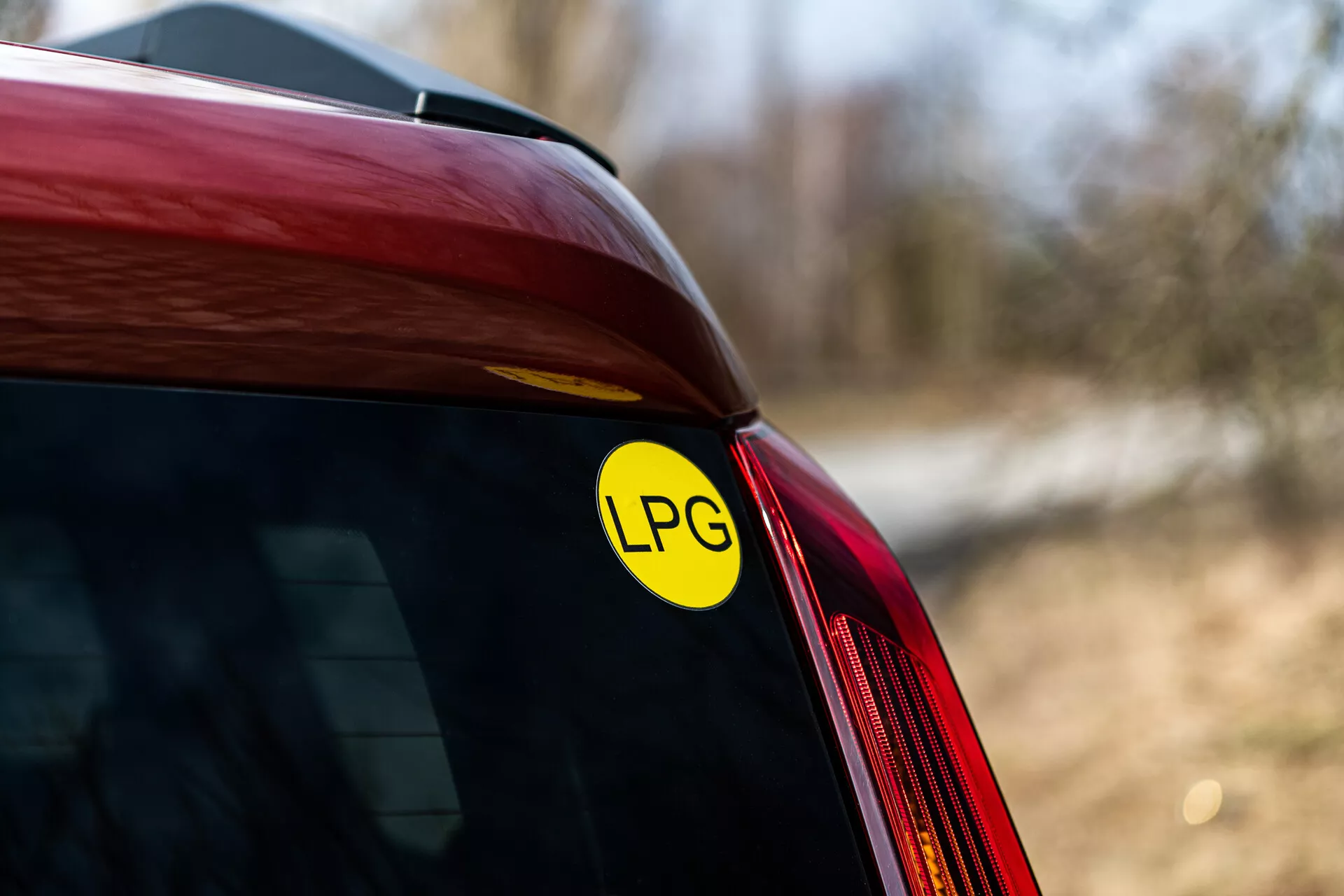 Detailný pohľad na zadný roh červeného Renault Arkana z roku 2021 s nálepkou LPG, ktorá označuje schopnosť vozidla používať skvapalnený ropný plyn, so zameraním na spojenie zadného okna a bočného panela, v prírodnom vonkajšom svetle